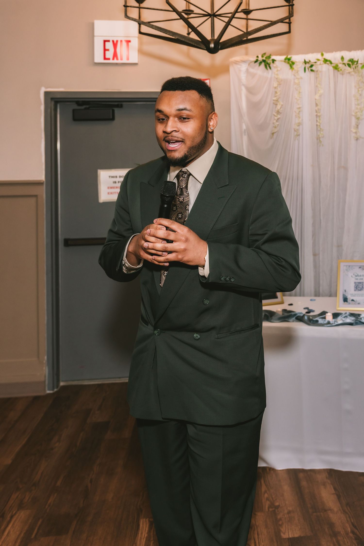 Man in dark green suit speaks into microphone at a wedding reception event.