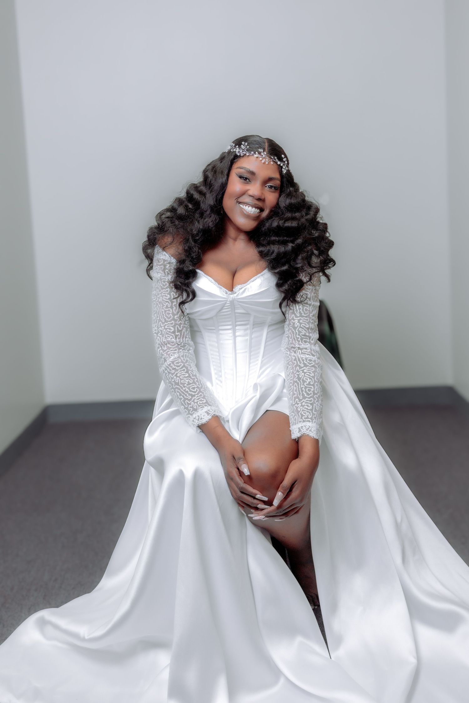 Smiling bride in a white corset wedding gown with lace sleeves and tiara seated gracefully.