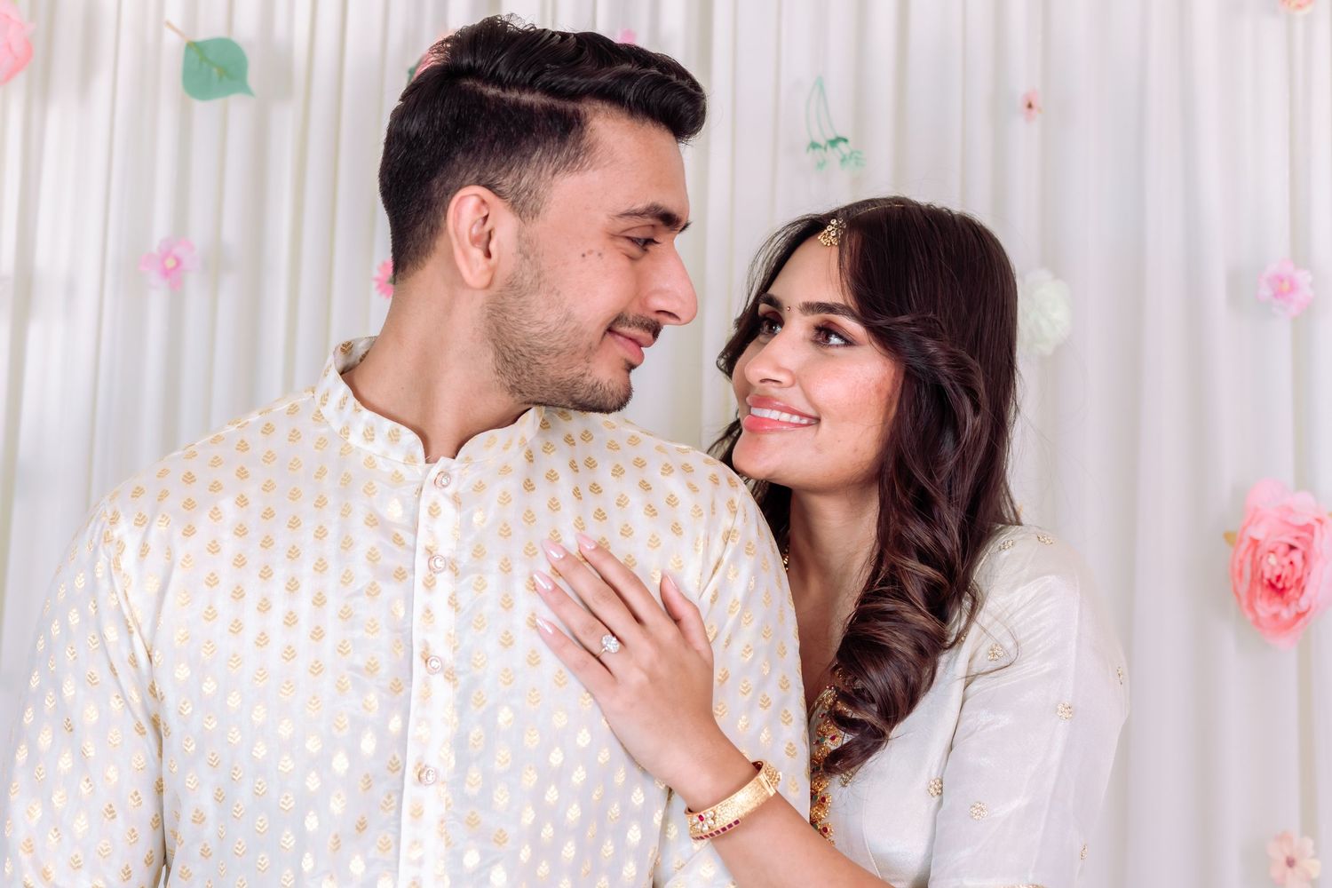 Indian couple in matching white outfits smiling at each other at engagement celebration.