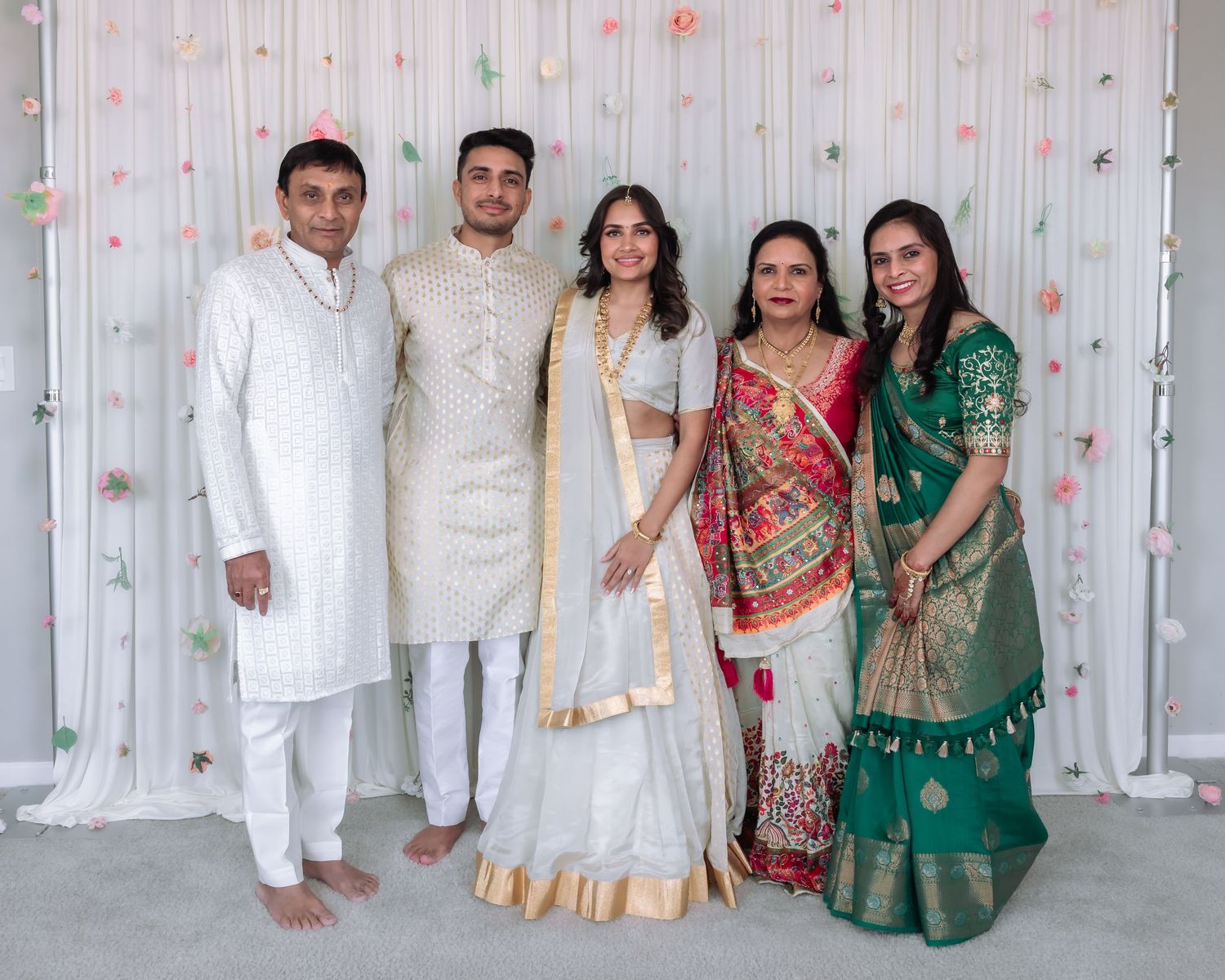 Five people in elegant Indian wedding outfits pose together in front of a floral white backdrop.