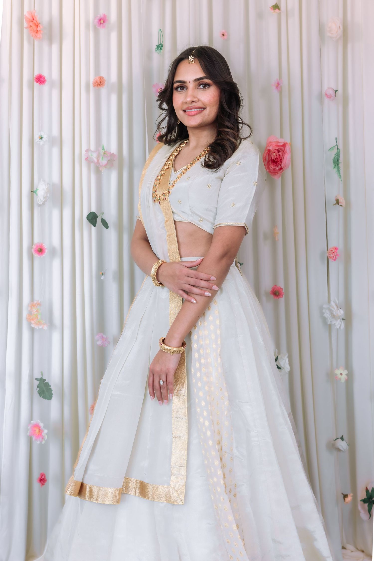 Young woman in white and gold lehenga smiling gracefully against a floral curtain backdrop.