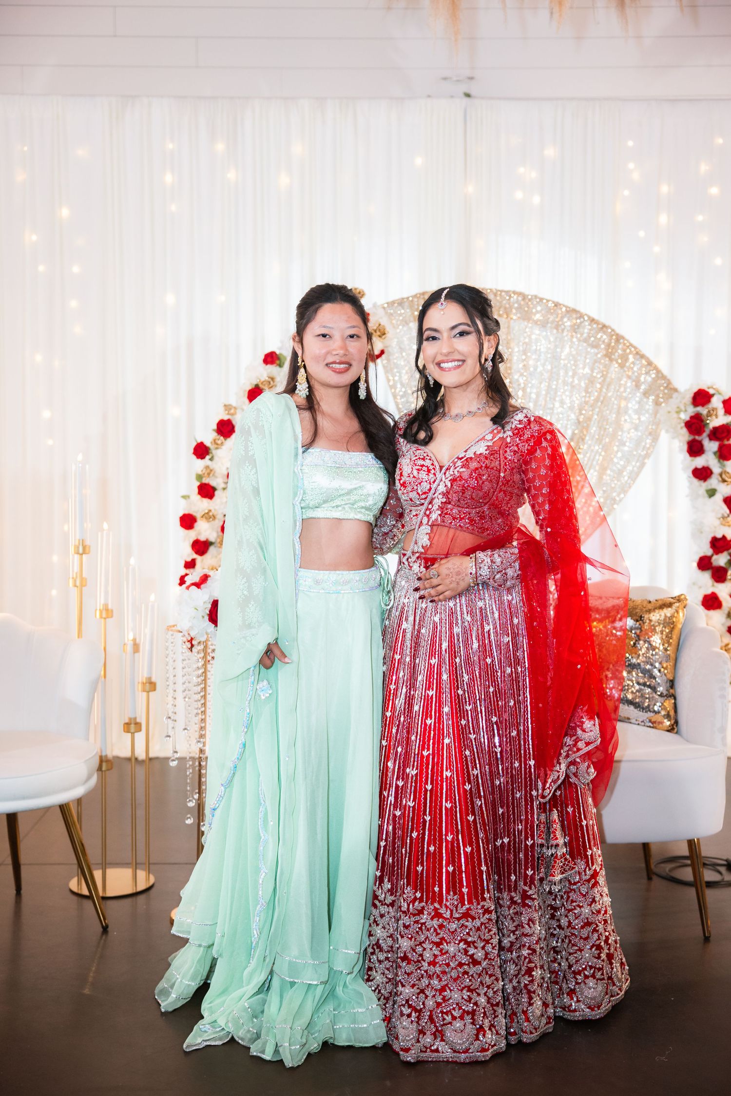 Two women posing in stunning Indian bridal outfits, one in red lehenga and one in mint green, at a decorated venue.