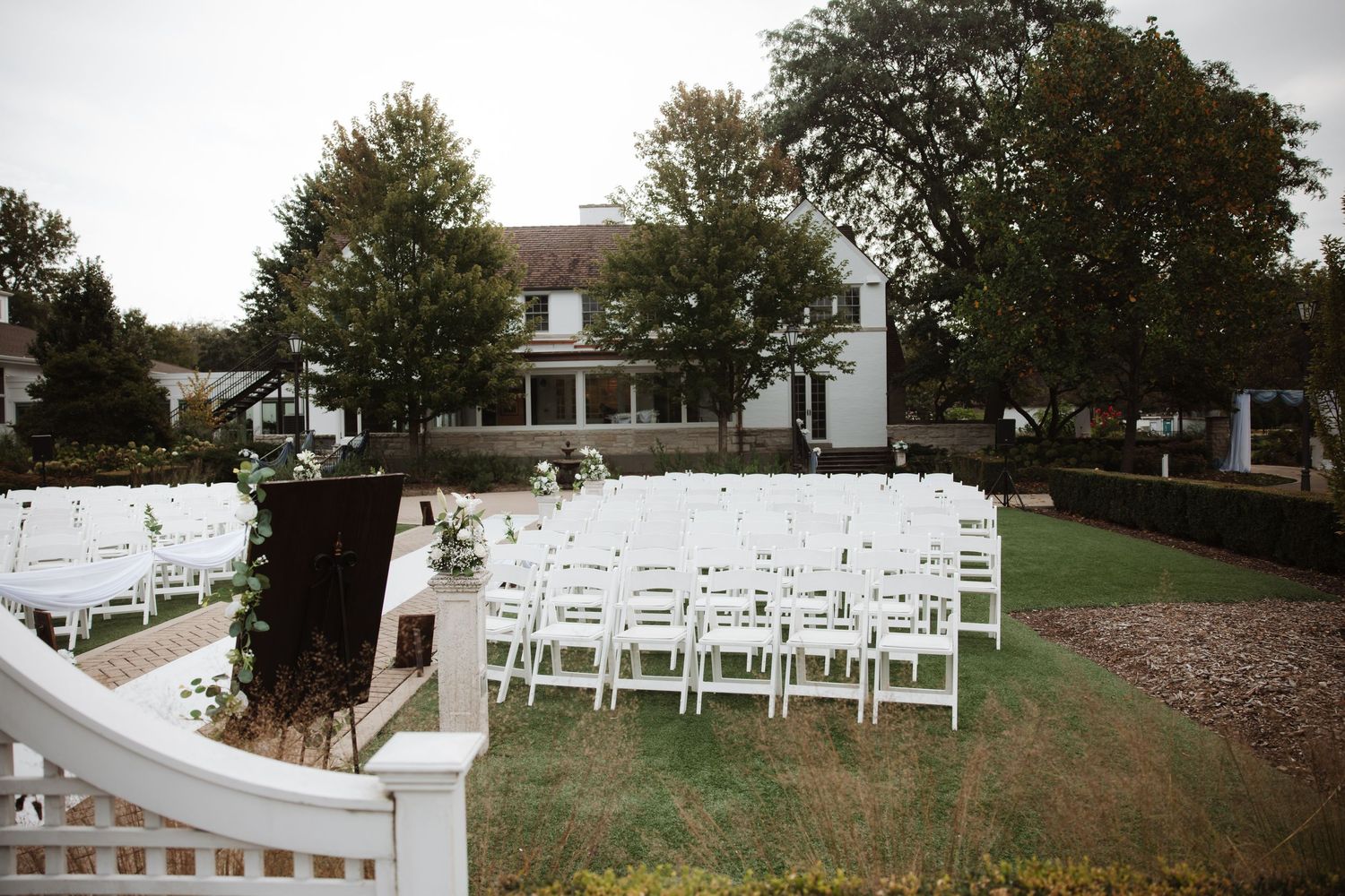 Outdoor wedding ceremony setup with white folding chairs on green lawn in front of white colonial manor house.