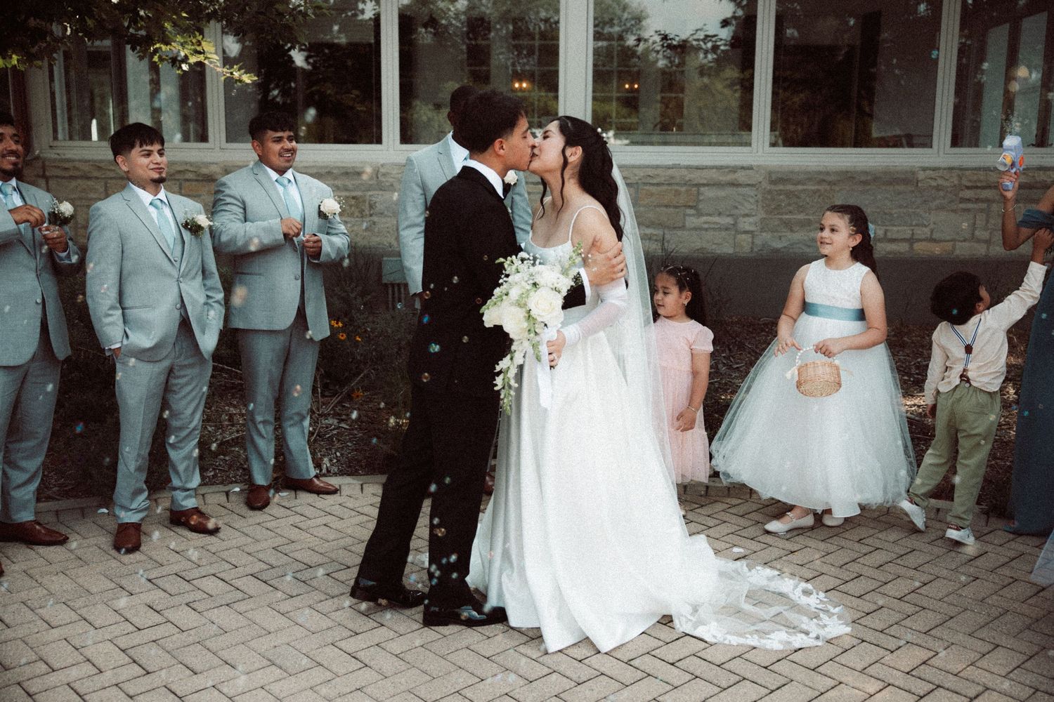 Bride and groom share a kiss surrounded by groomsmen in light blue suits and flower girls at outdoor wedding ceremony.