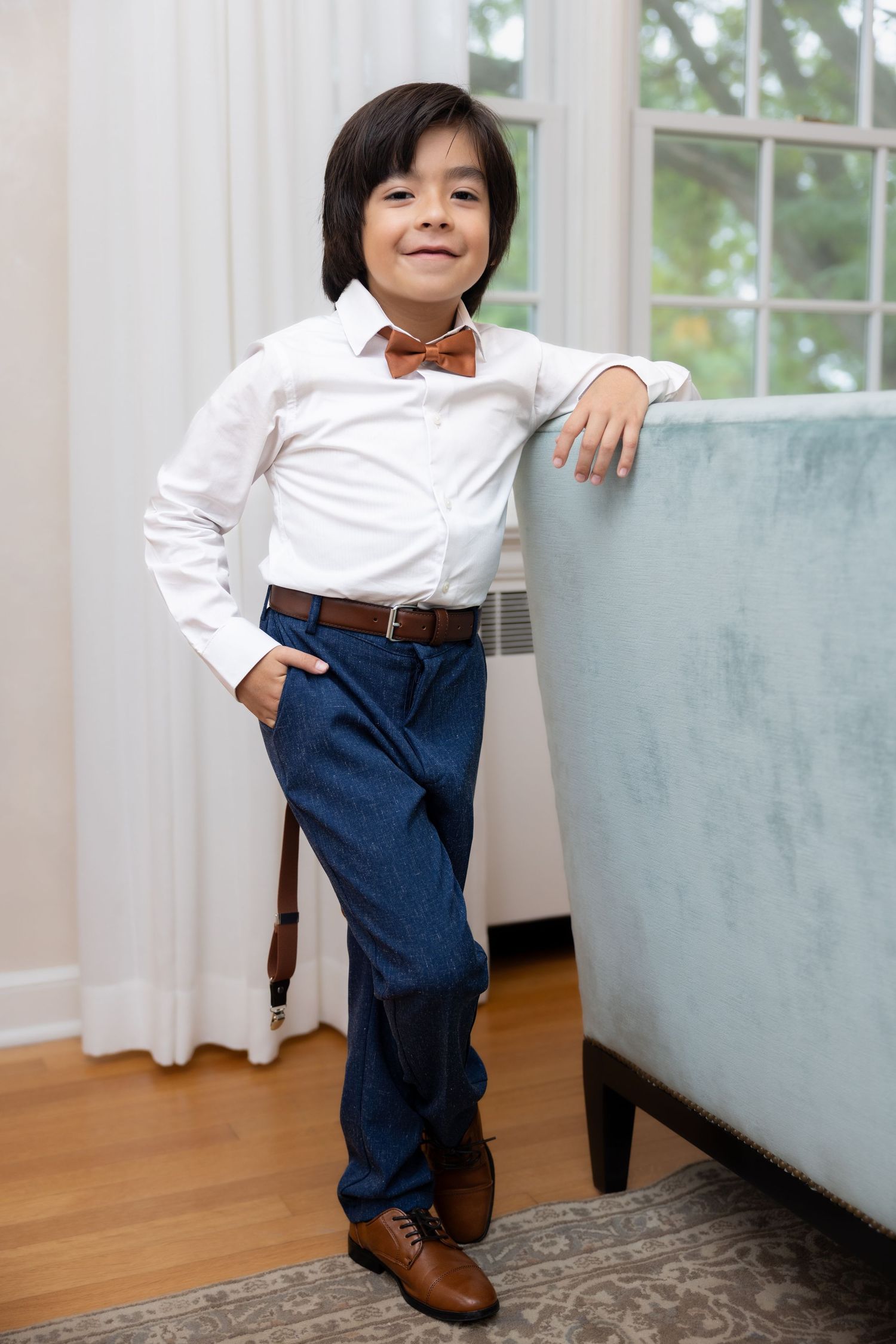 Dapper young boy in white shirt, blue trousers, and brown bow tie smiling by a couch.