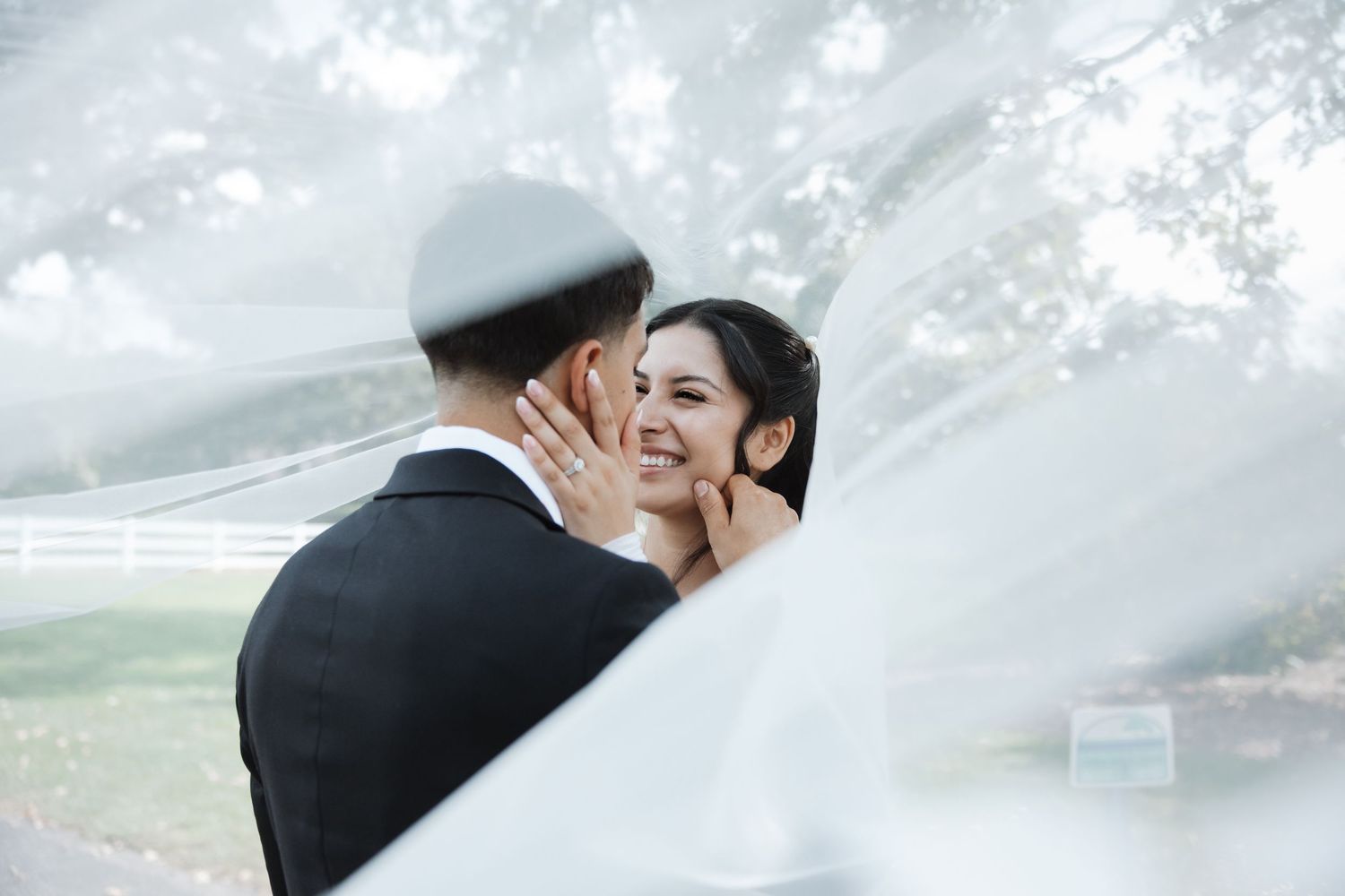 Joyful bride smiles at groom under a flowing white veil with trees and white fence in background.