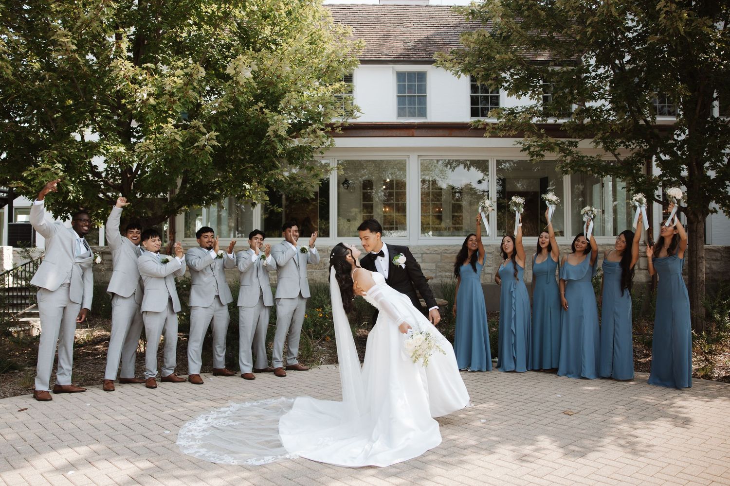 Groom dips bride as wedding party cheers with arms raised outside elegant white building.
