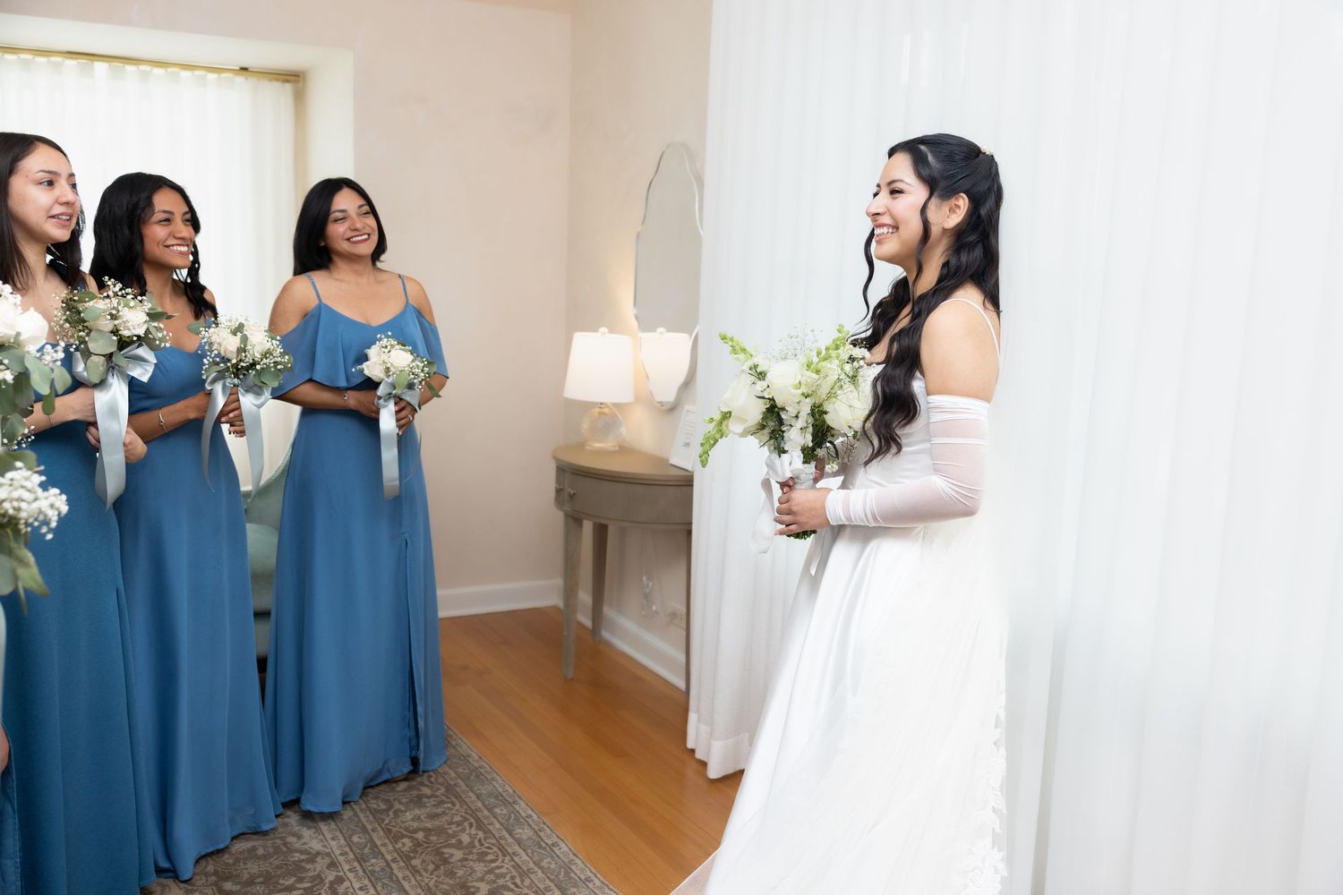 Bride in white gown smiles at bridesmaids in blue dresses holding white bouquets during wedding preparations.