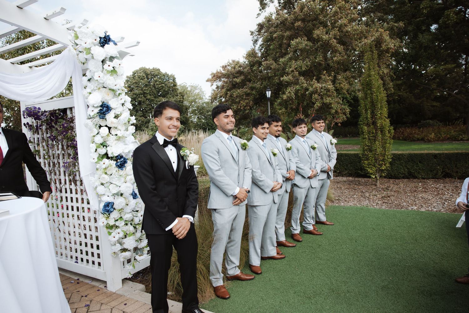 Groom in black tuxedo stands at altar with groomsmen in gray suits at outdoor wedding.