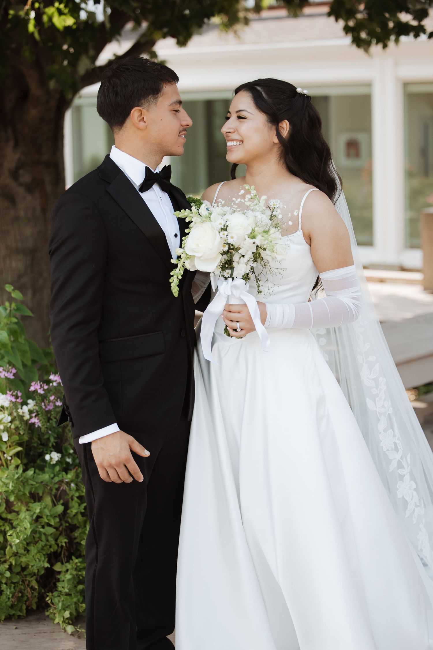 Smiling bride and groom in formal wedding attire gaze at each other outdoors among garden greenery.