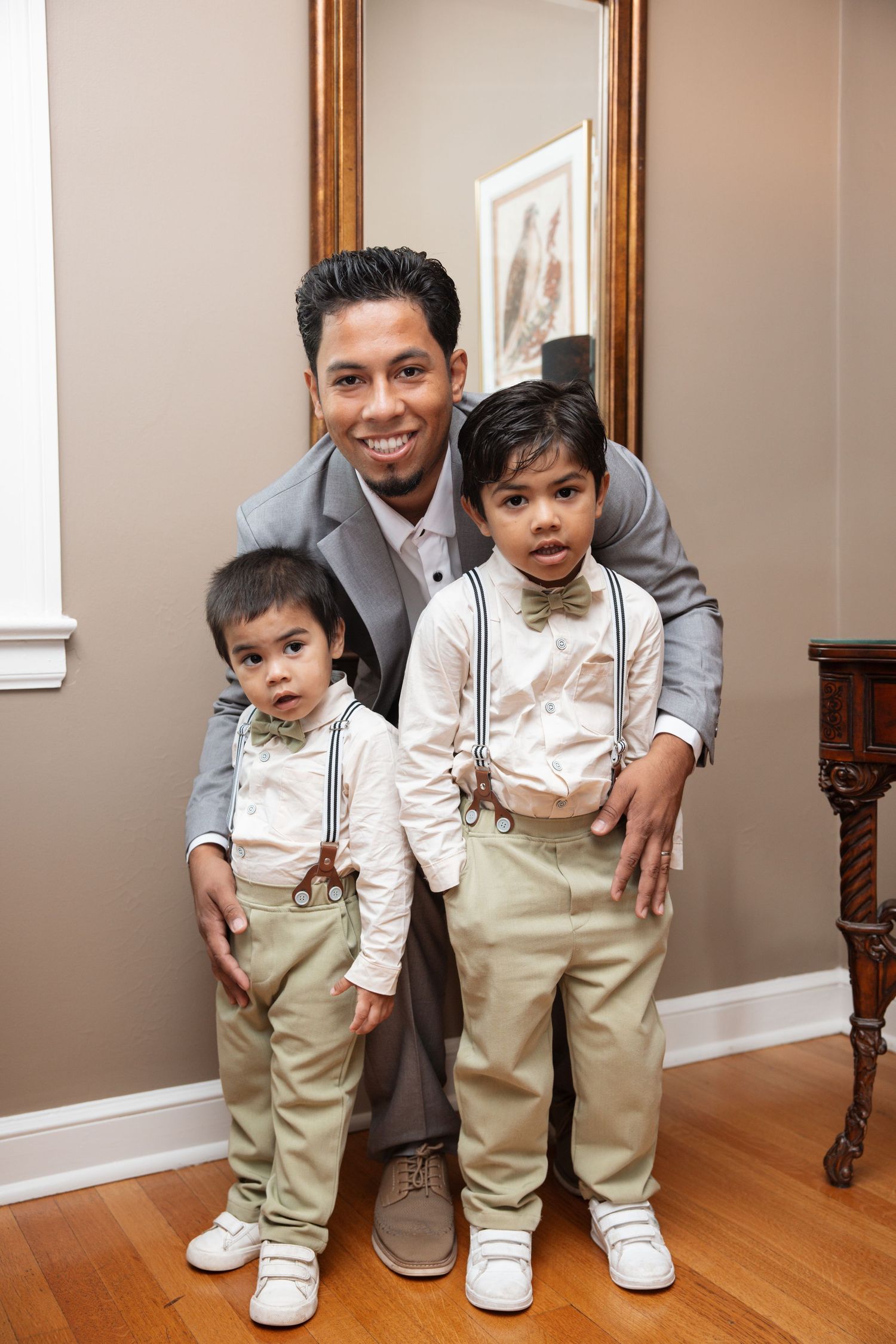 Man in gray suit posing with two small boys wearing bow ties and suspenders before a large mirror.
