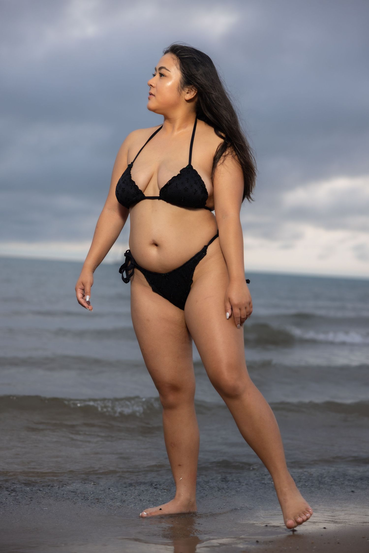 Curvy woman in black bikini standing confidently on beach shoreline with overcast sky and ocean waves.