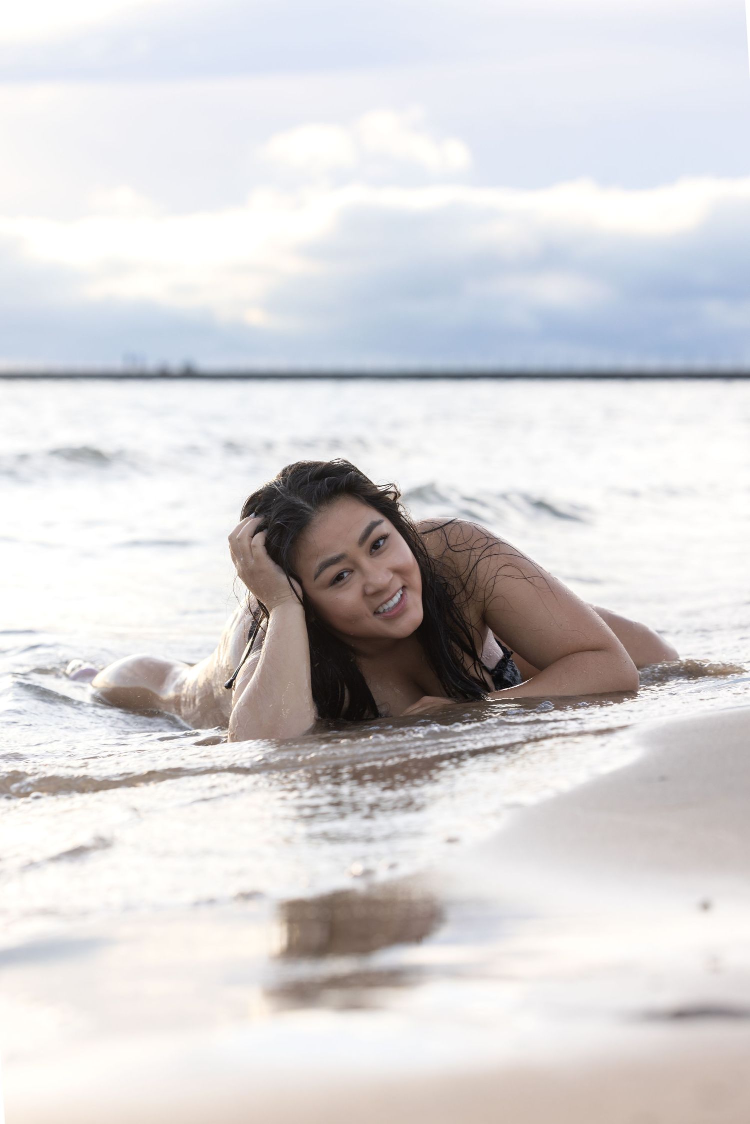 Smiling woman with wet hair lying in shallow ocean waves on a cloudy beach.