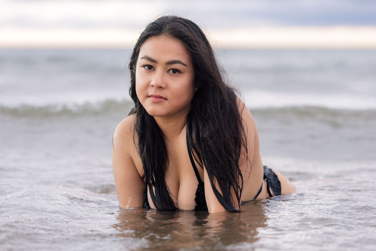 Woman with long dark hair in black bikini posing in shallow ocean water on overcast day.