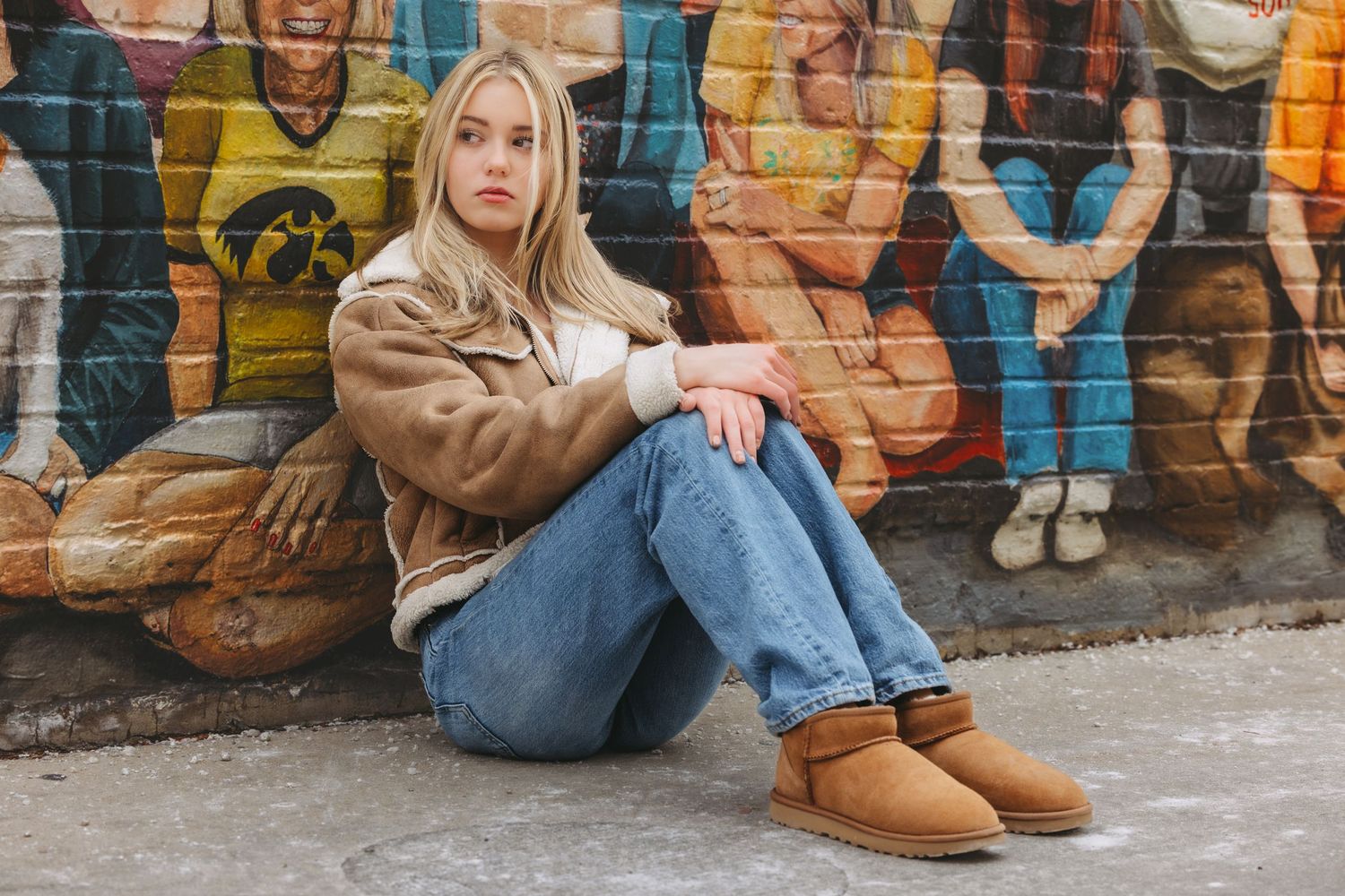 A blonde woman in a tan jacket, jeans, and UGG boots sits against a vibrant outdoor mural wall.