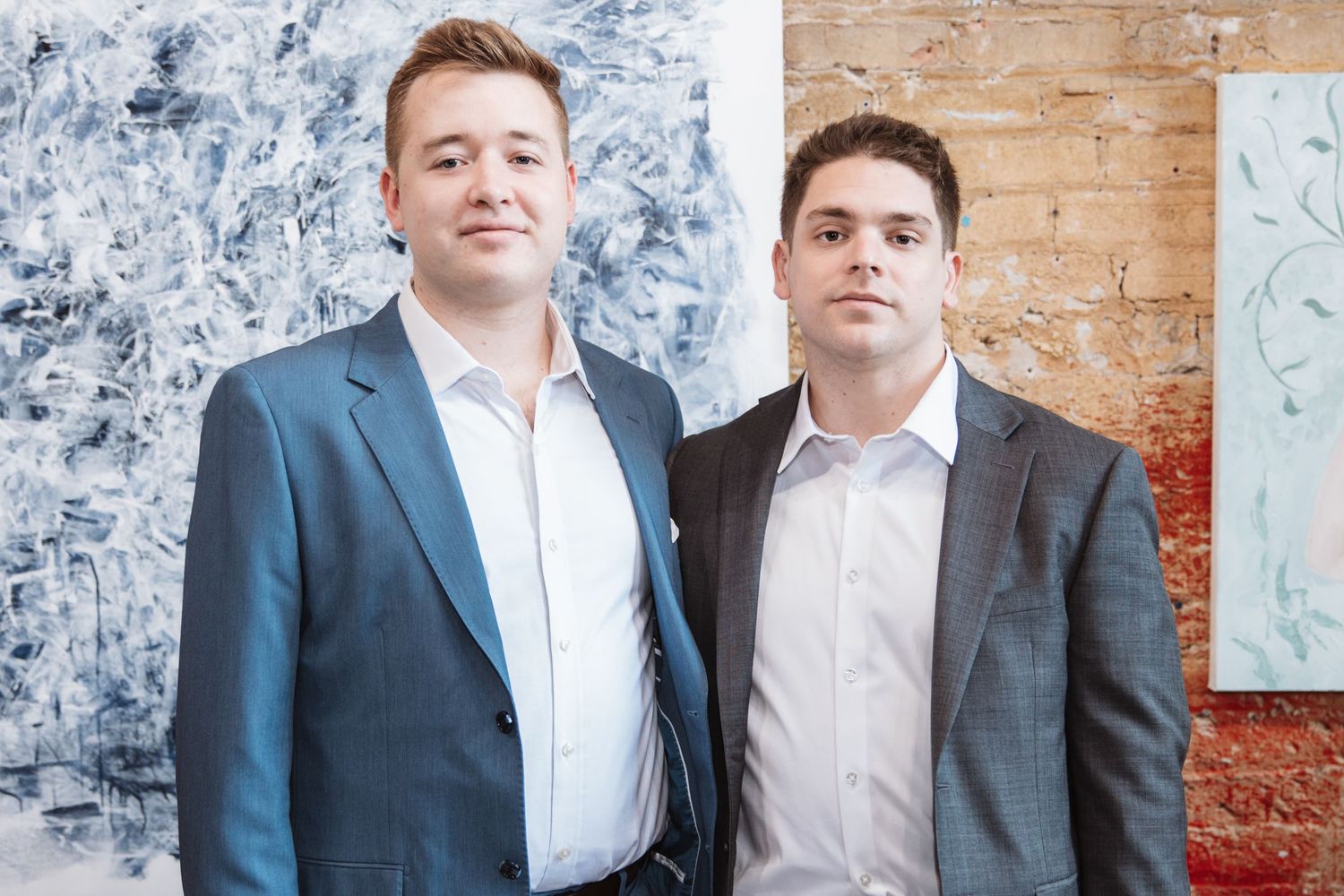 Two well-dressed men in suits smile together in front of abstract artwork at an art gallery.