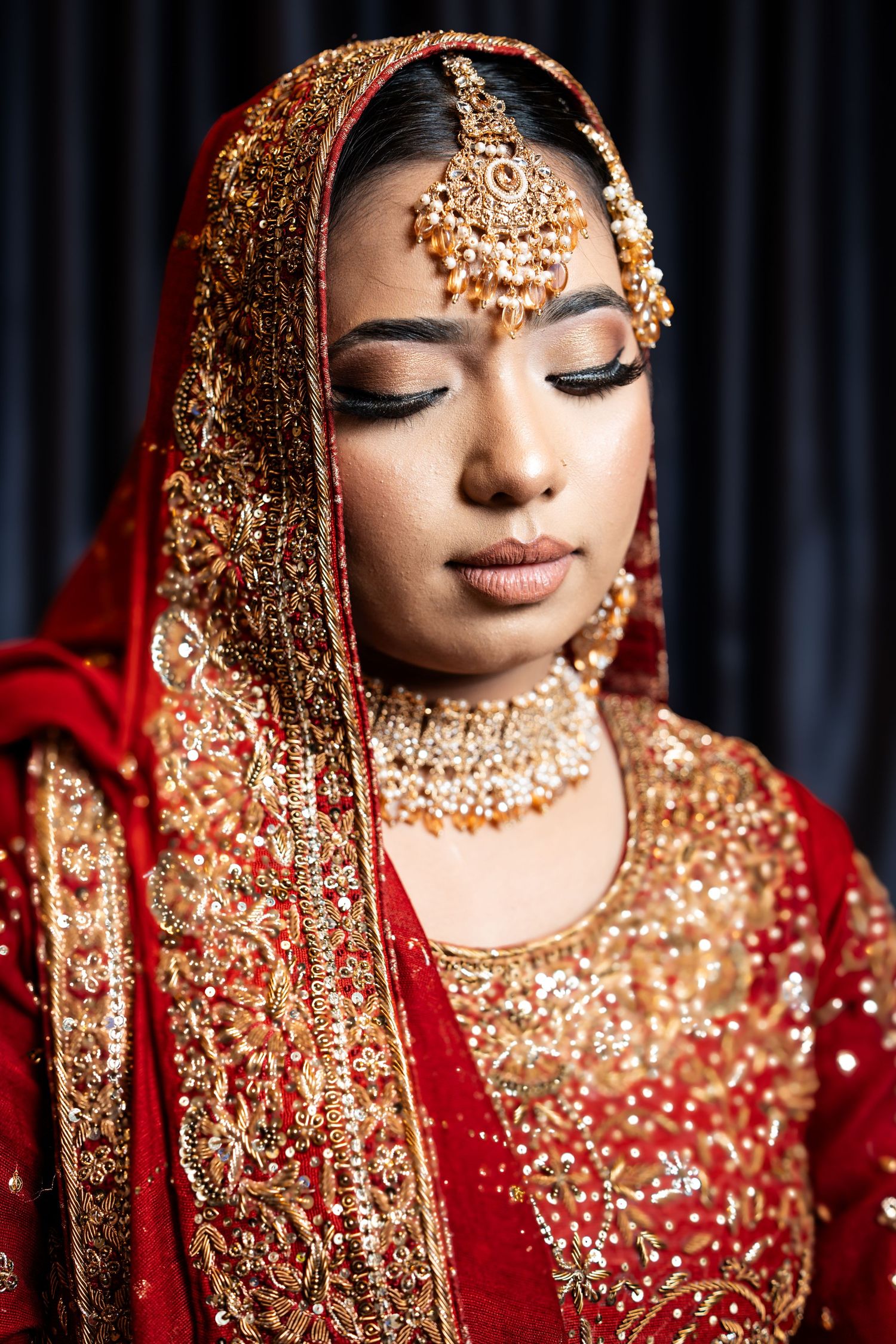 Portrait of a bride in red and gold Indian bridal attire and jewelry with downcast eyes.