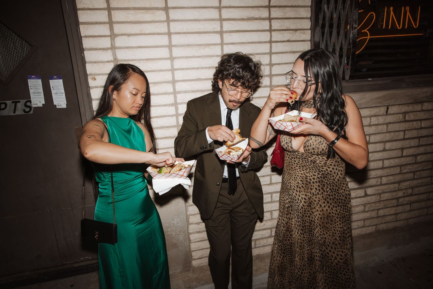 Three wedding guests in formal attire eating takeout food outside a venue entrance at night.