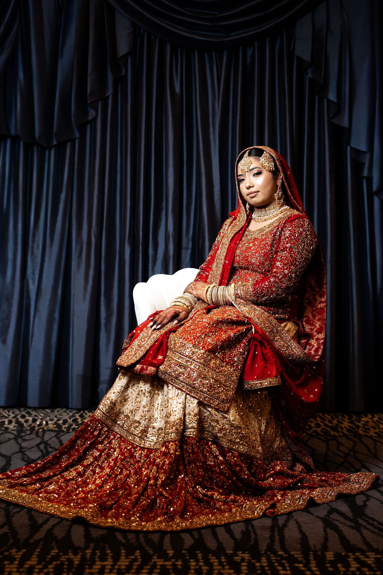 Bride in red and gold South Asian bridal lehenga seated regally against blue drapes.