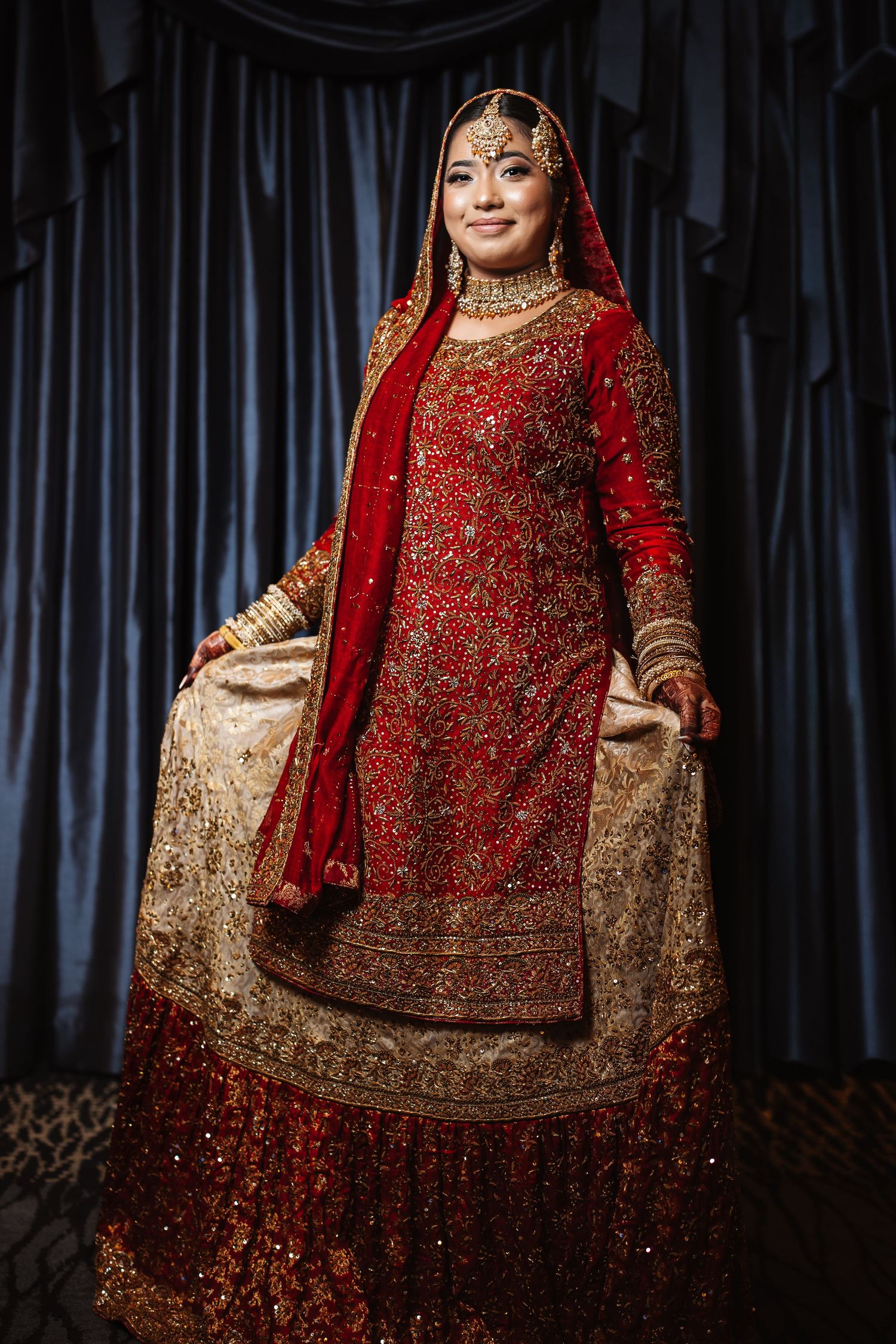 Smiling South Asian bride in ornate red and gold bridal lehenga with dupatta and traditional jewelry.