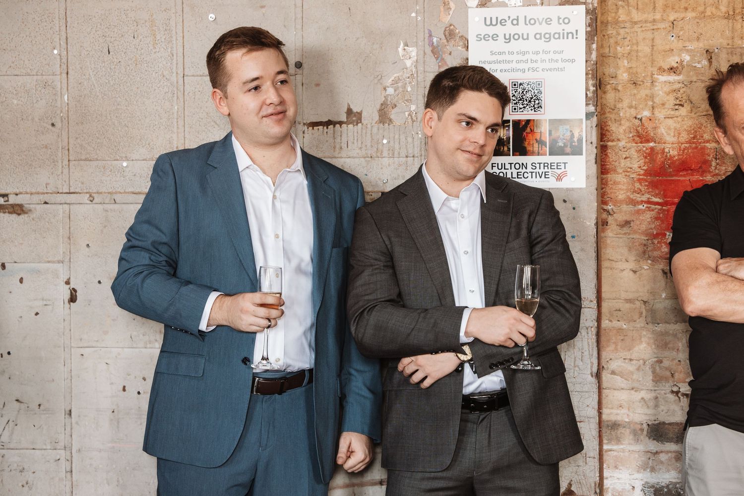 Two men in suits smiling and holding champagne glasses at an art gallery opening event.