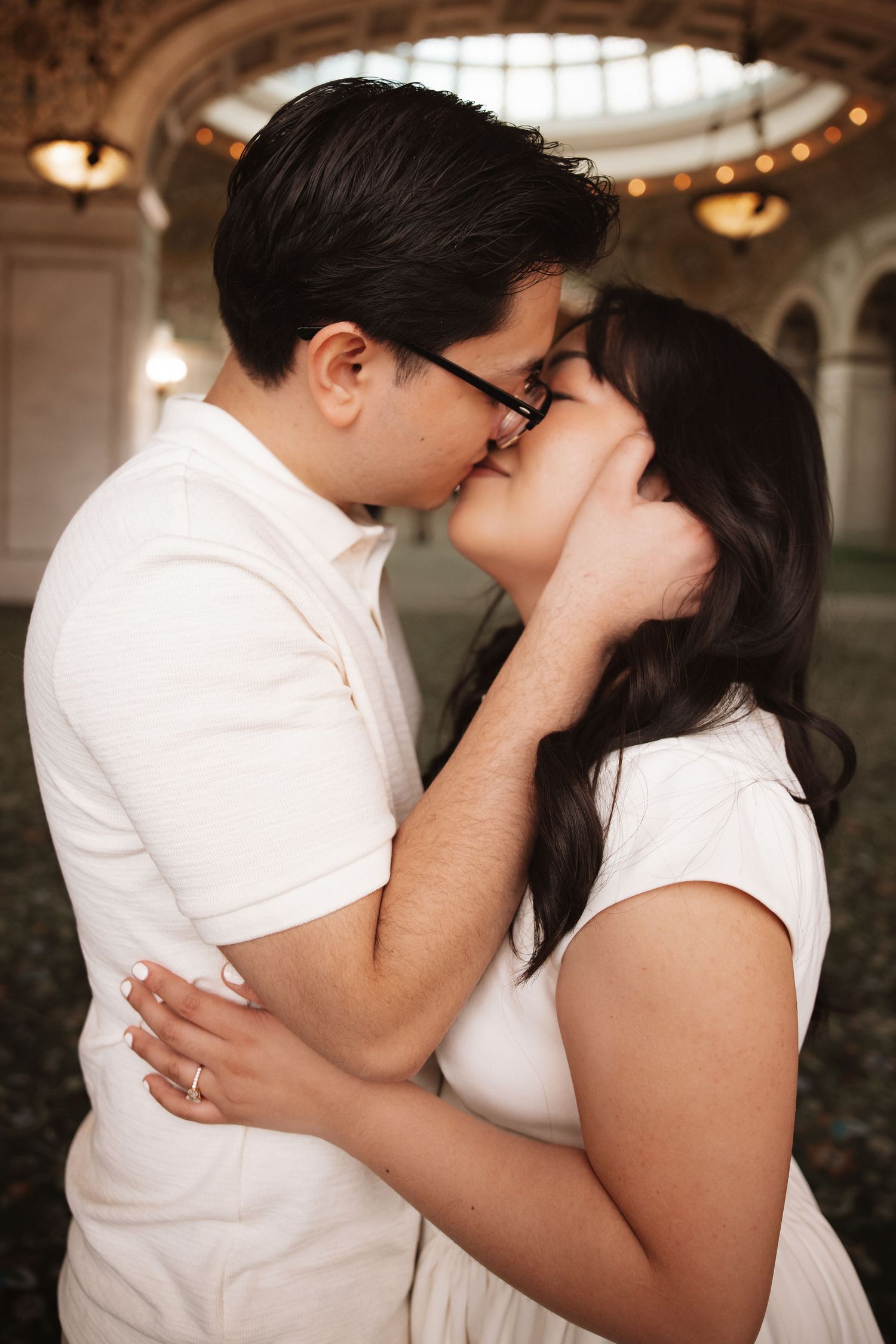 Engaged couple sharing a passionate kiss in ornate historic building with warm chandelier bokeh background.