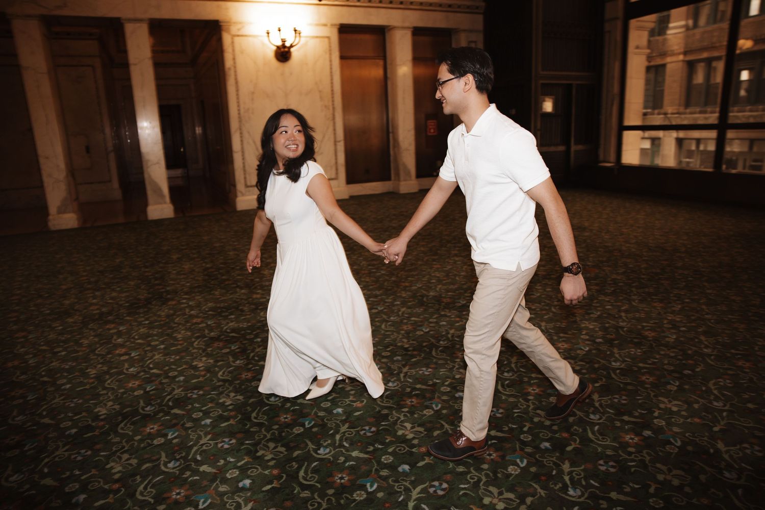 Bride and groom hold hands walking across ornate carpeted ballroom.