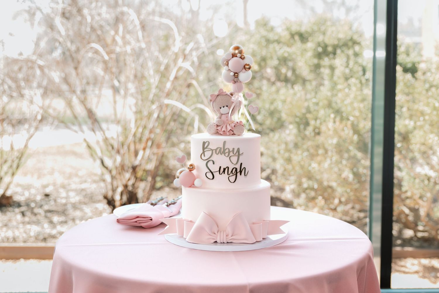 Elegant two-tier Baby Singh baby shower cake with teddy bear topper on a pink tablecloth.