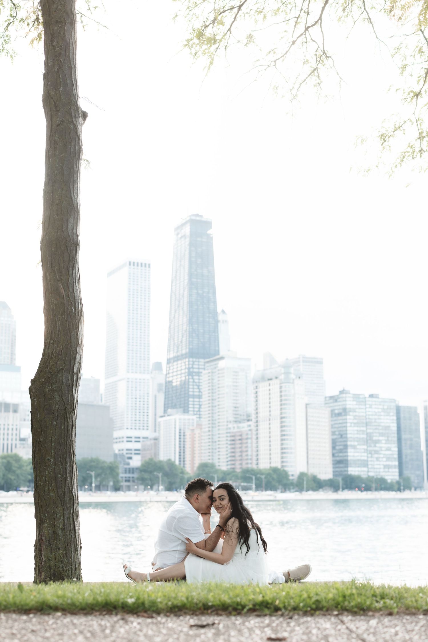 Couple sitting together on grass by lakefront with Chicago city skyline in background near a tall tree.