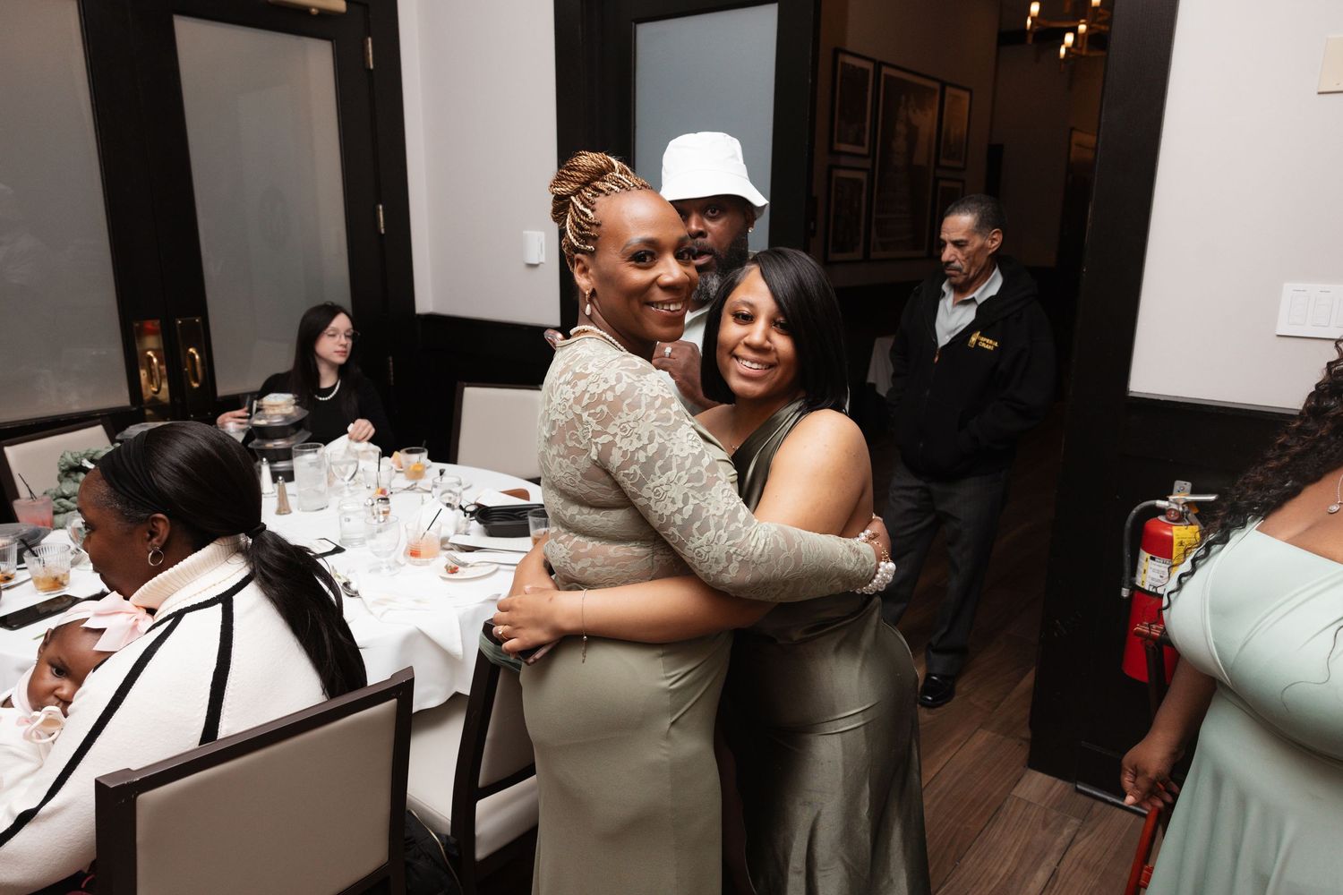 Two women sharing a warm hug at a wedding reception dinner, guests and decorated tables visible in background.