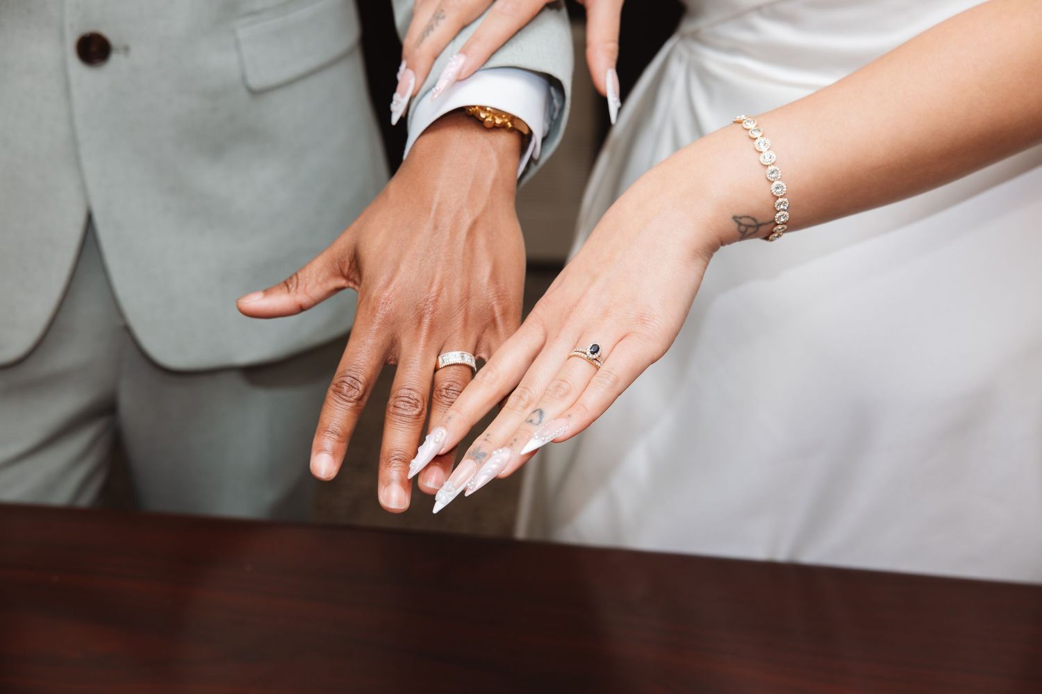 Newlywed couple displaying wedding rings on their hands, bride in white dress with bracelet, groom in gray suit.