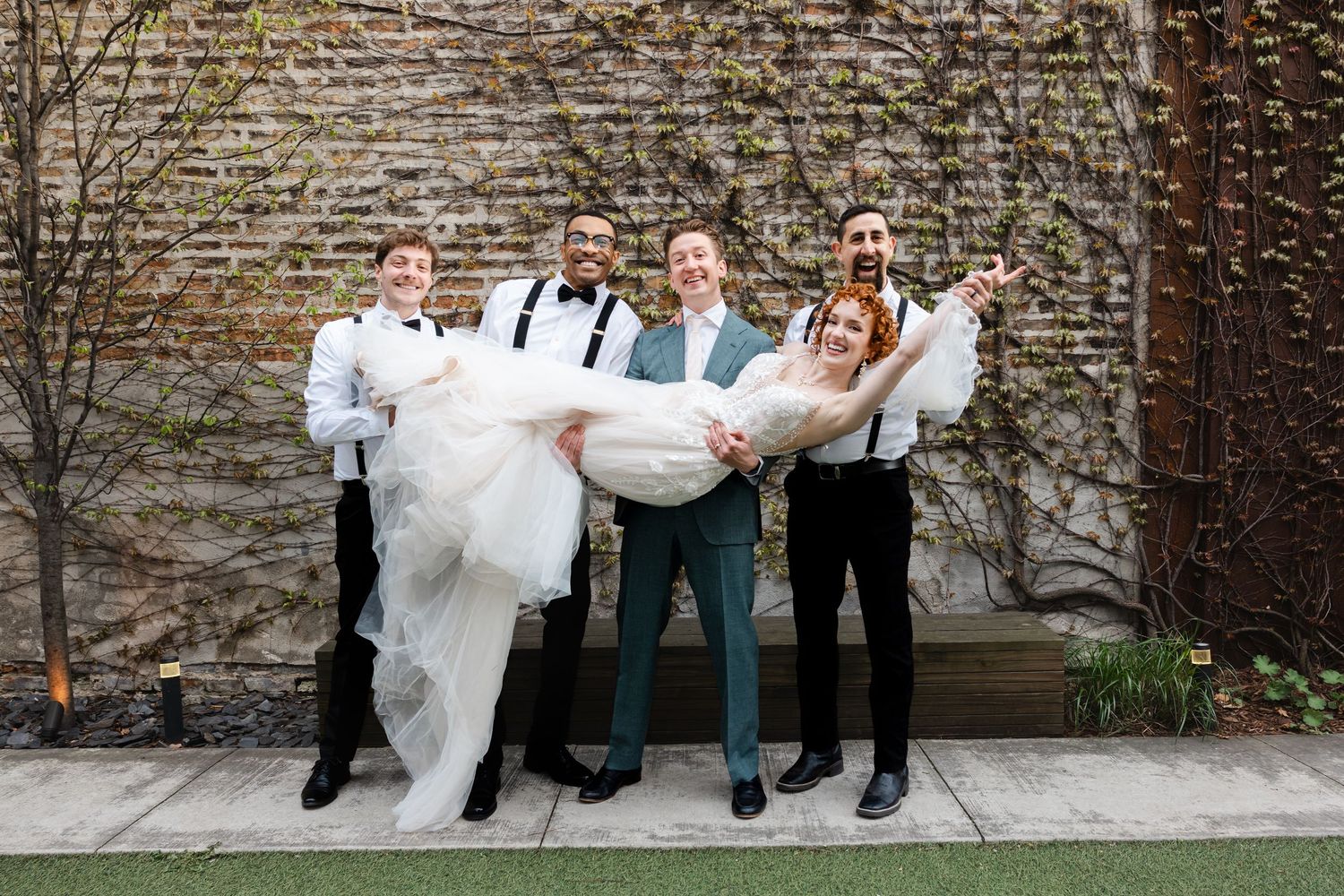 Groomsmen in suspenders playfully lift smiling bride while groom stands center at outdoor wedding.