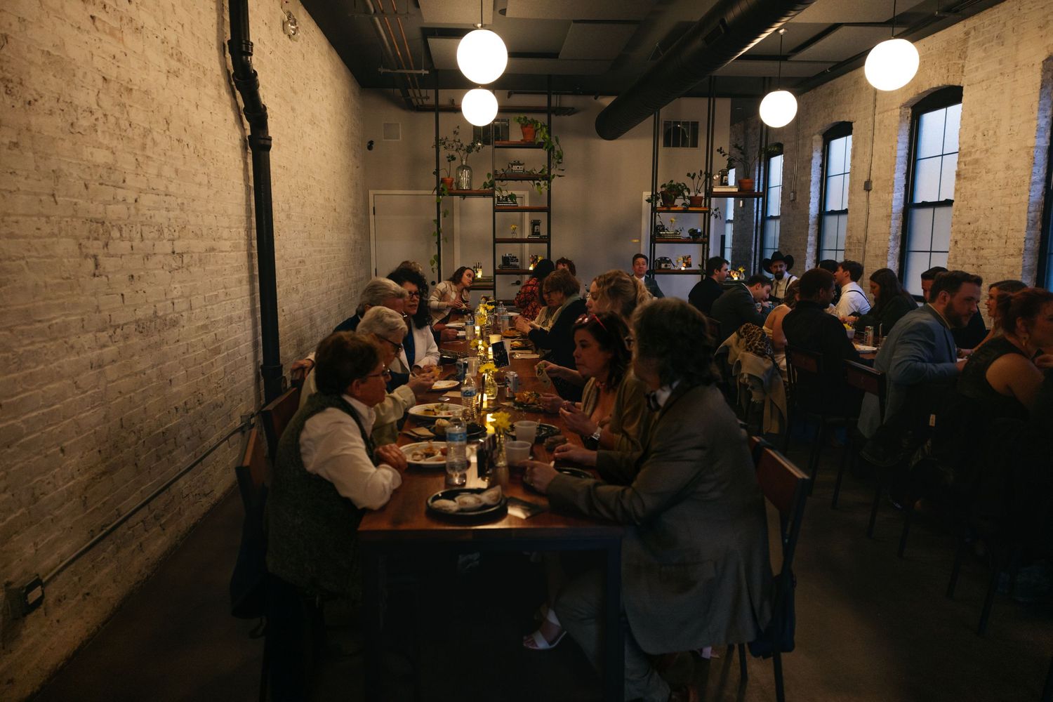 Guests dining at long tables in a rustic industrial venue with exposed brick walls and globe pendant lights.