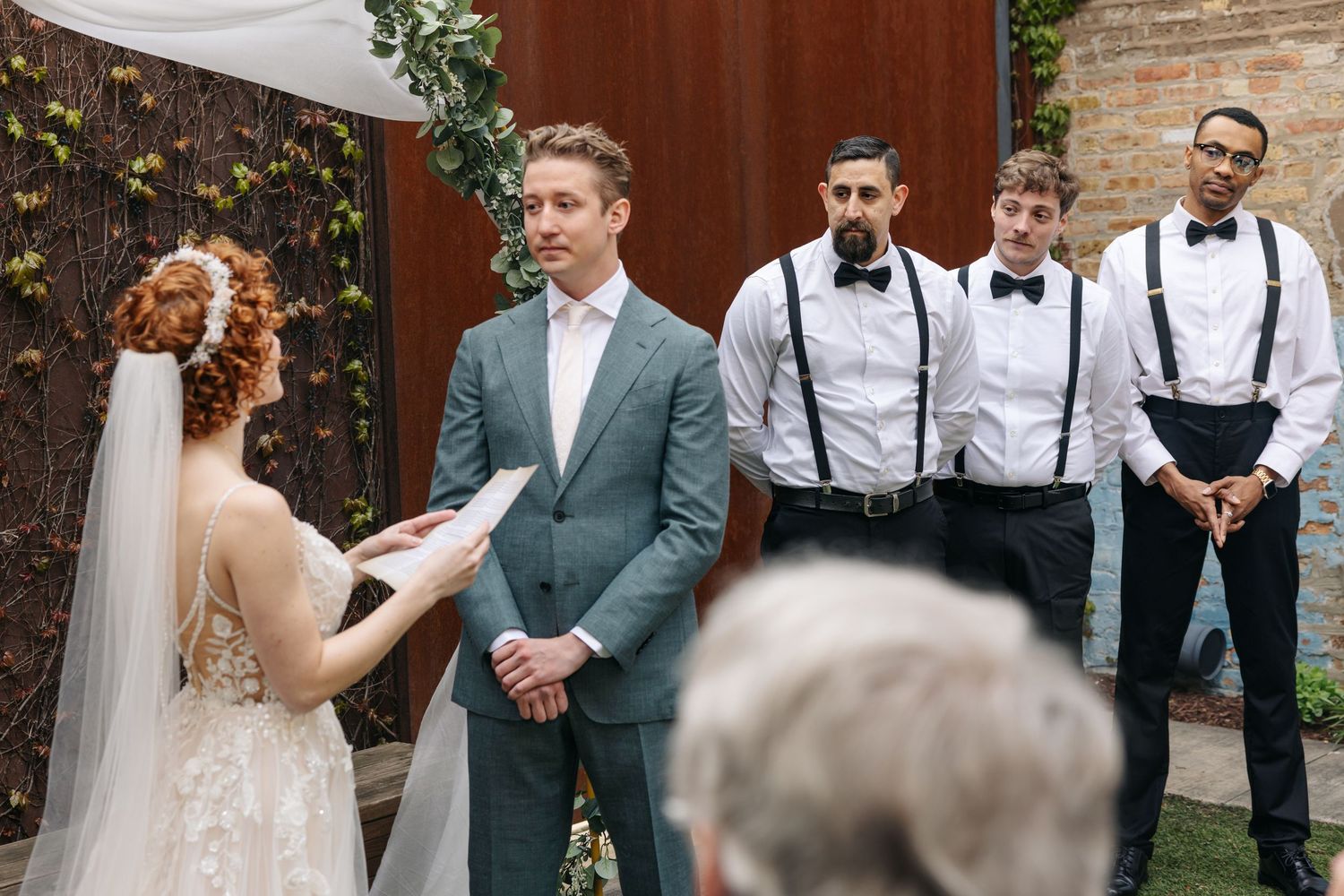 Bride and groom exchange vows outdoors, with groomsmen in suspenders and bow ties standing nearby.