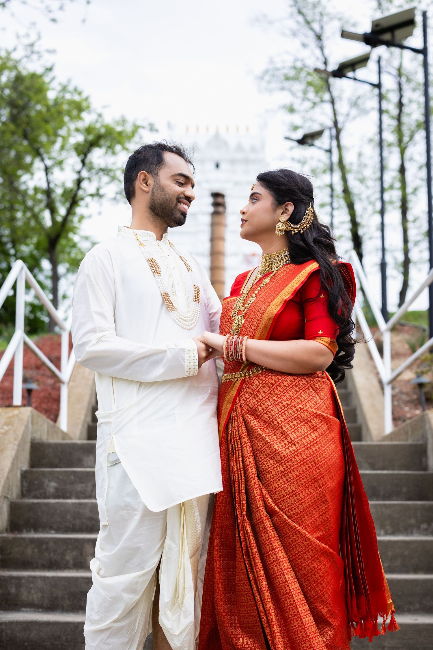Indian couple in traditional wedding attire, man in white kurta and woman in red saree with gold jewelry, standing on steps.