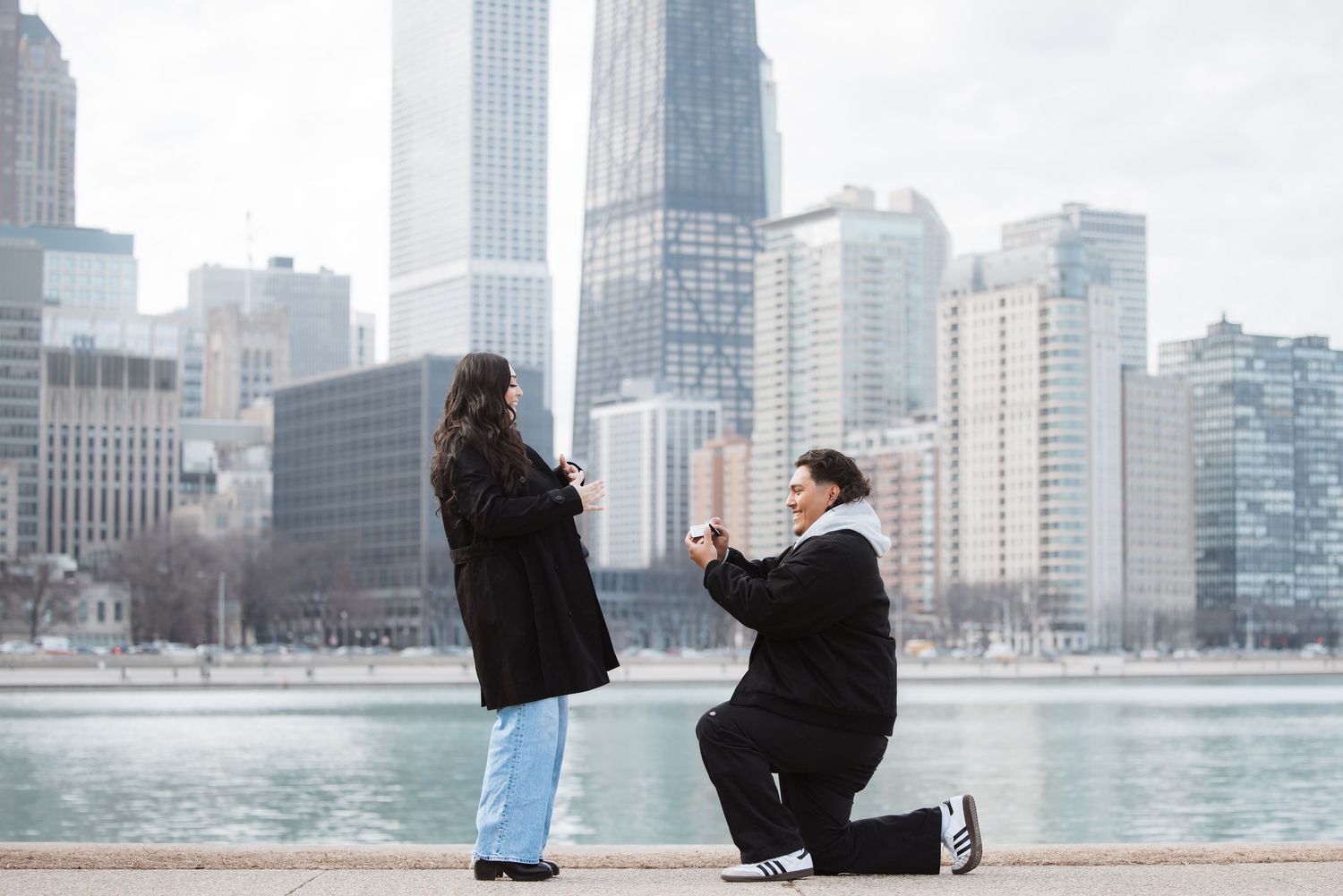 Man proposes on one knee to woman by Chicago lakefront with city skyline in background.