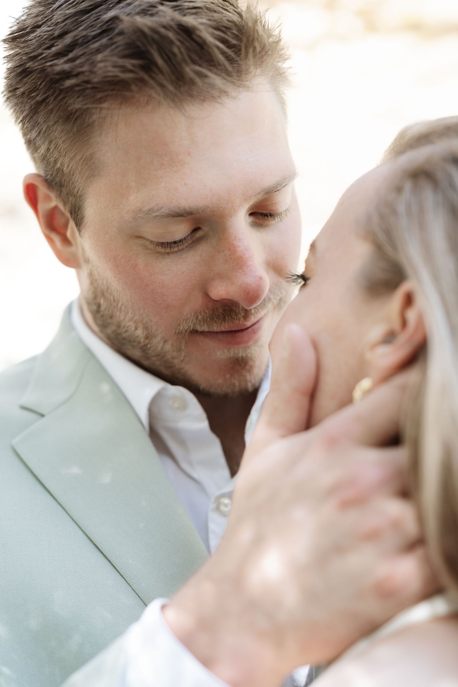 Close-up of groom in light suit gently cradling bride's face with tender expression outdoors.