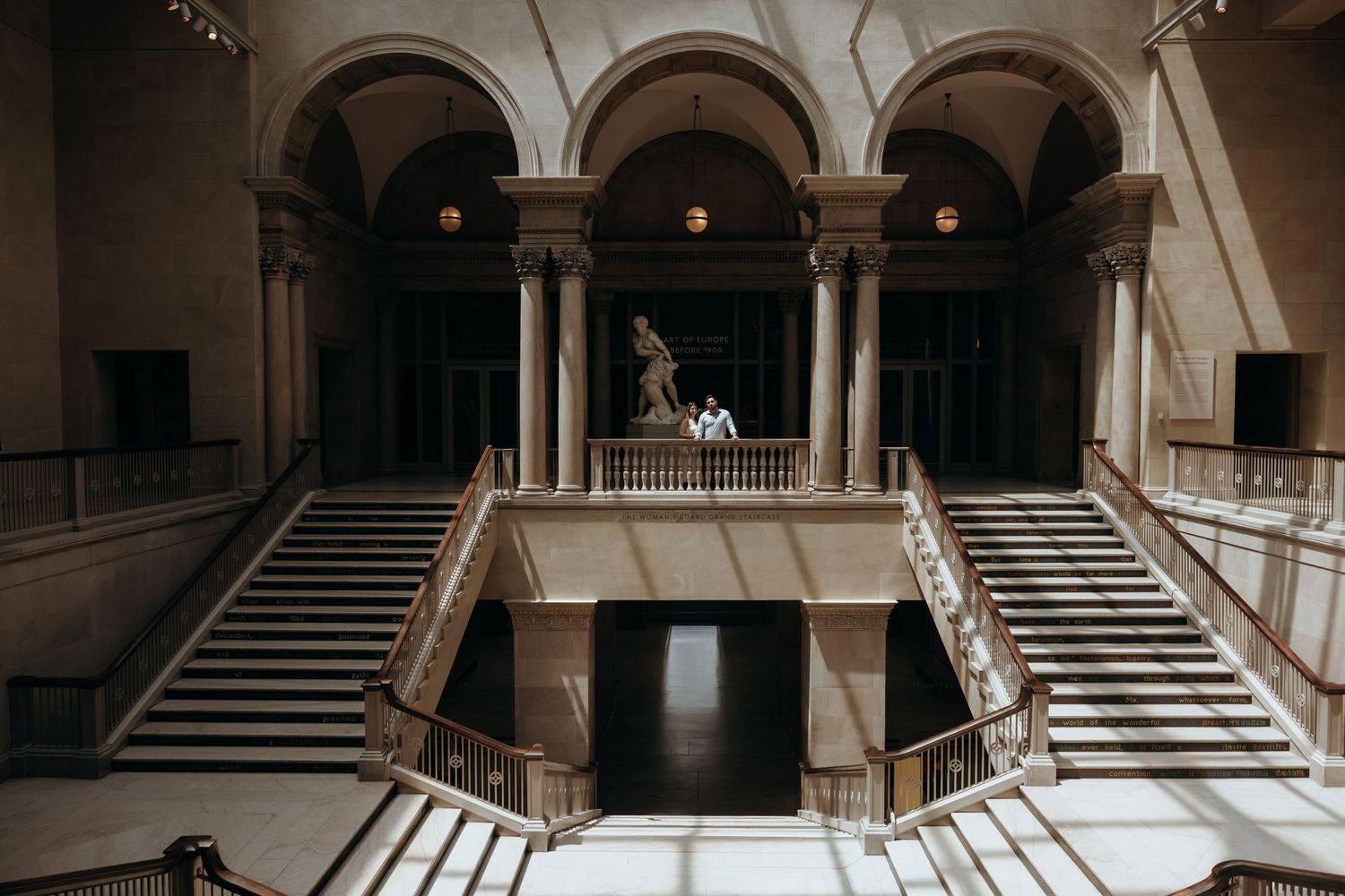 Grand neoclassical museum interior with sweeping staircases, arched columns, and a bronze sculpture centerpiece.