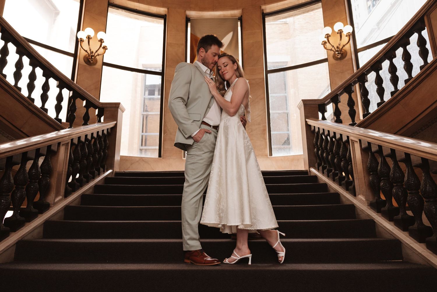 Bride and groom share a romantic embrace on an elegant staircase in a grand venue.