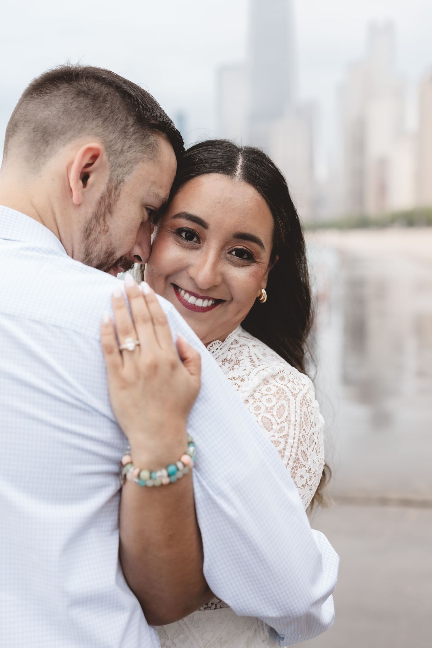 Smiling bride in white lace dress hugging groom outdoors, showing engagement ring, city skyline in background.