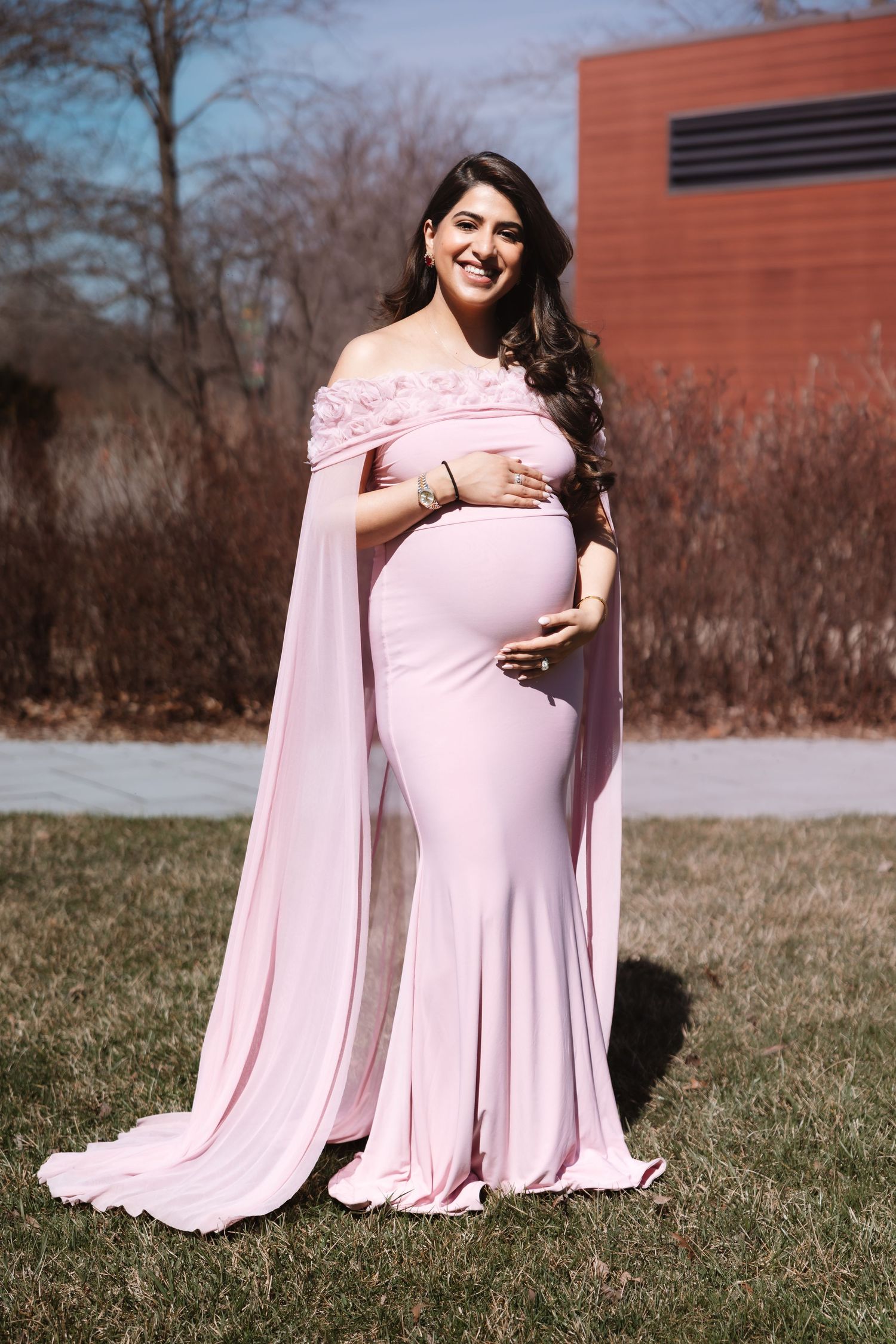 Radiant pregnant woman in pink cape maternity gown smiles outdoors against natural backdrop.