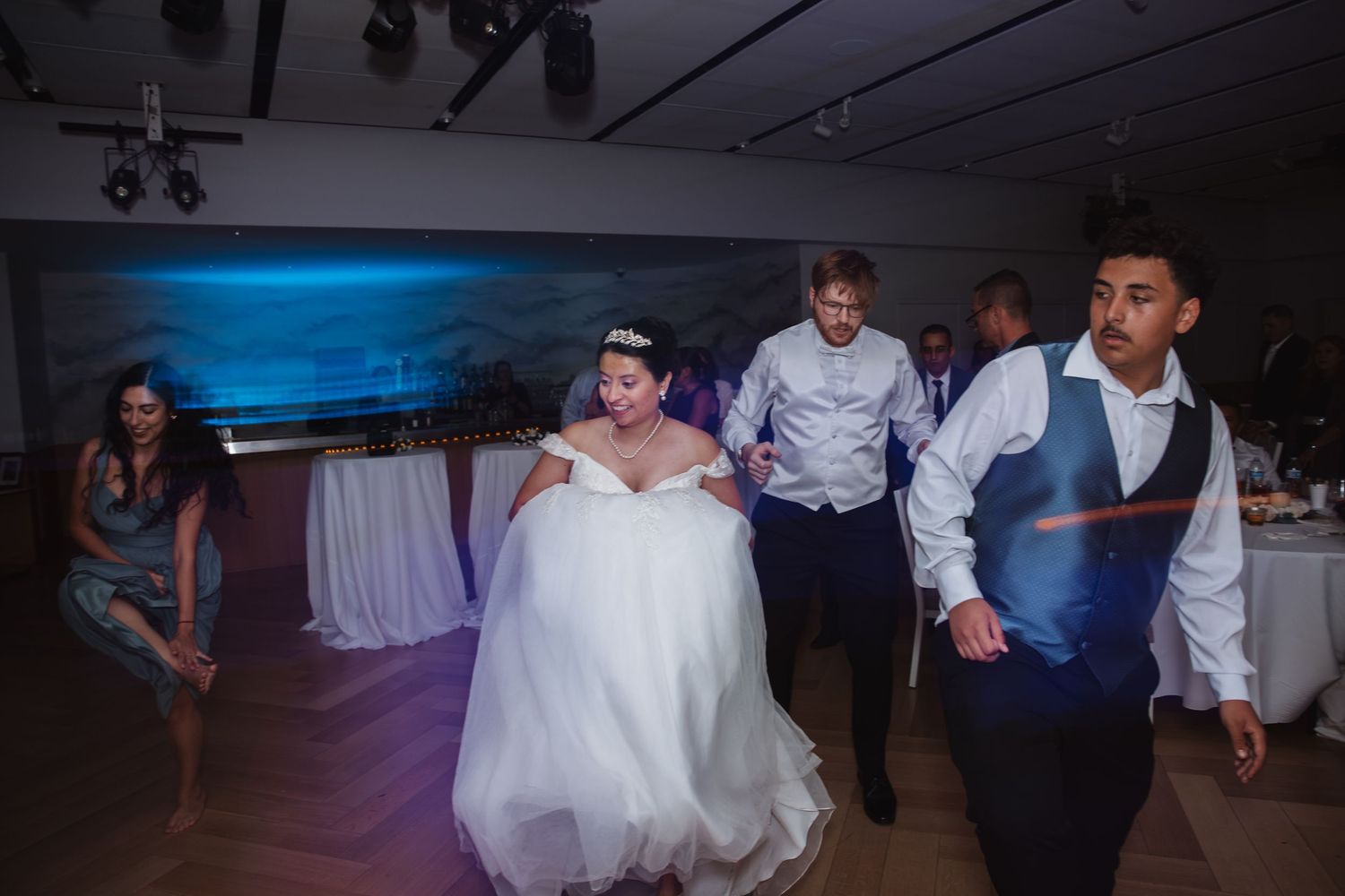 Bride in white gown dancing energetically with guests at wedding reception.