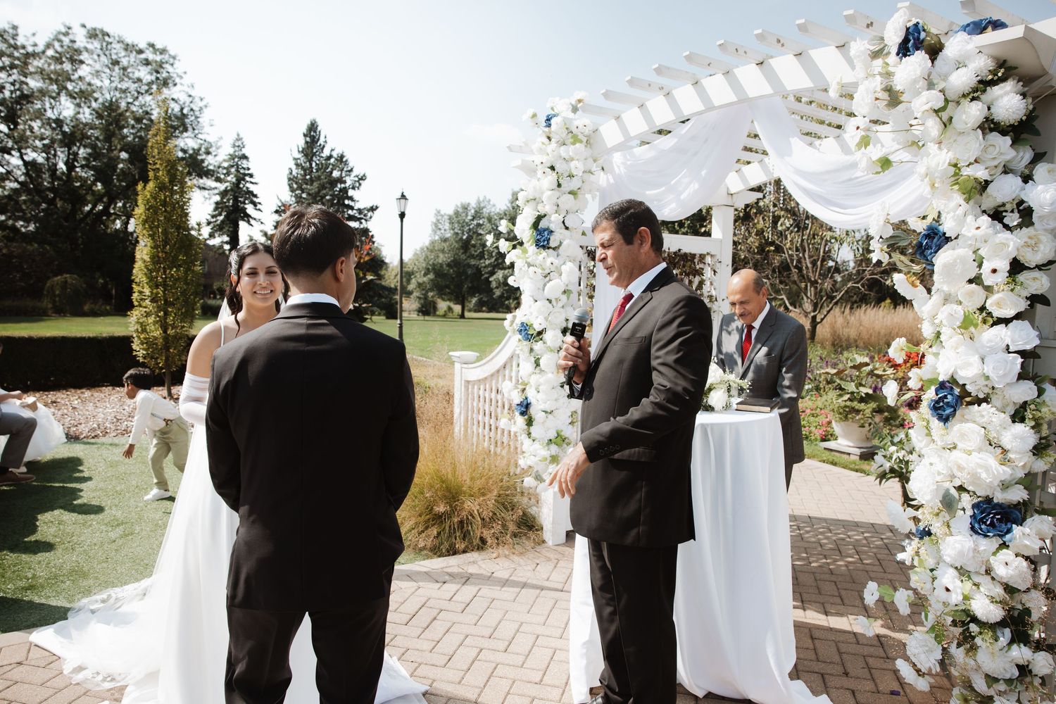 Officiant leads bride and groom in vows under white floral arch at outdoor wedding.