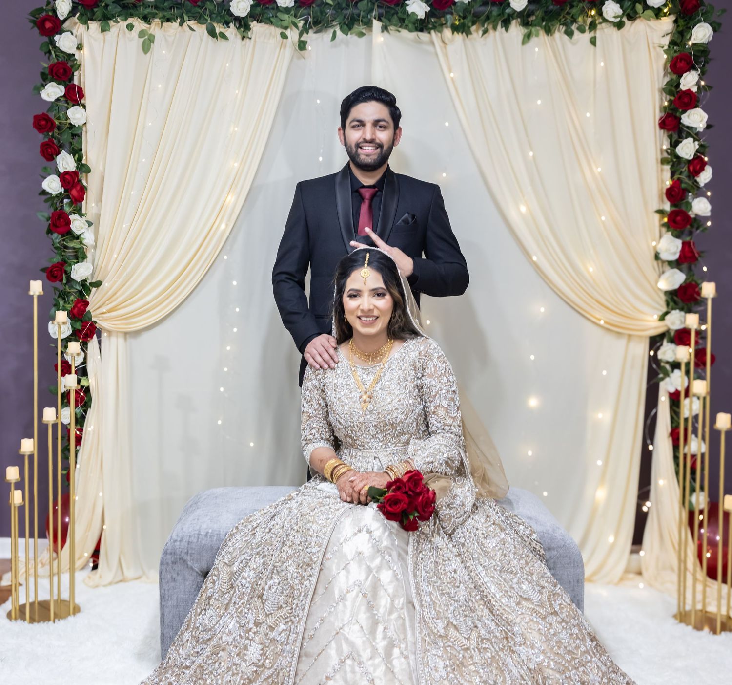 South Asian bride in gold lehenga holds red roses as groom stands behind her at decorated mandap.