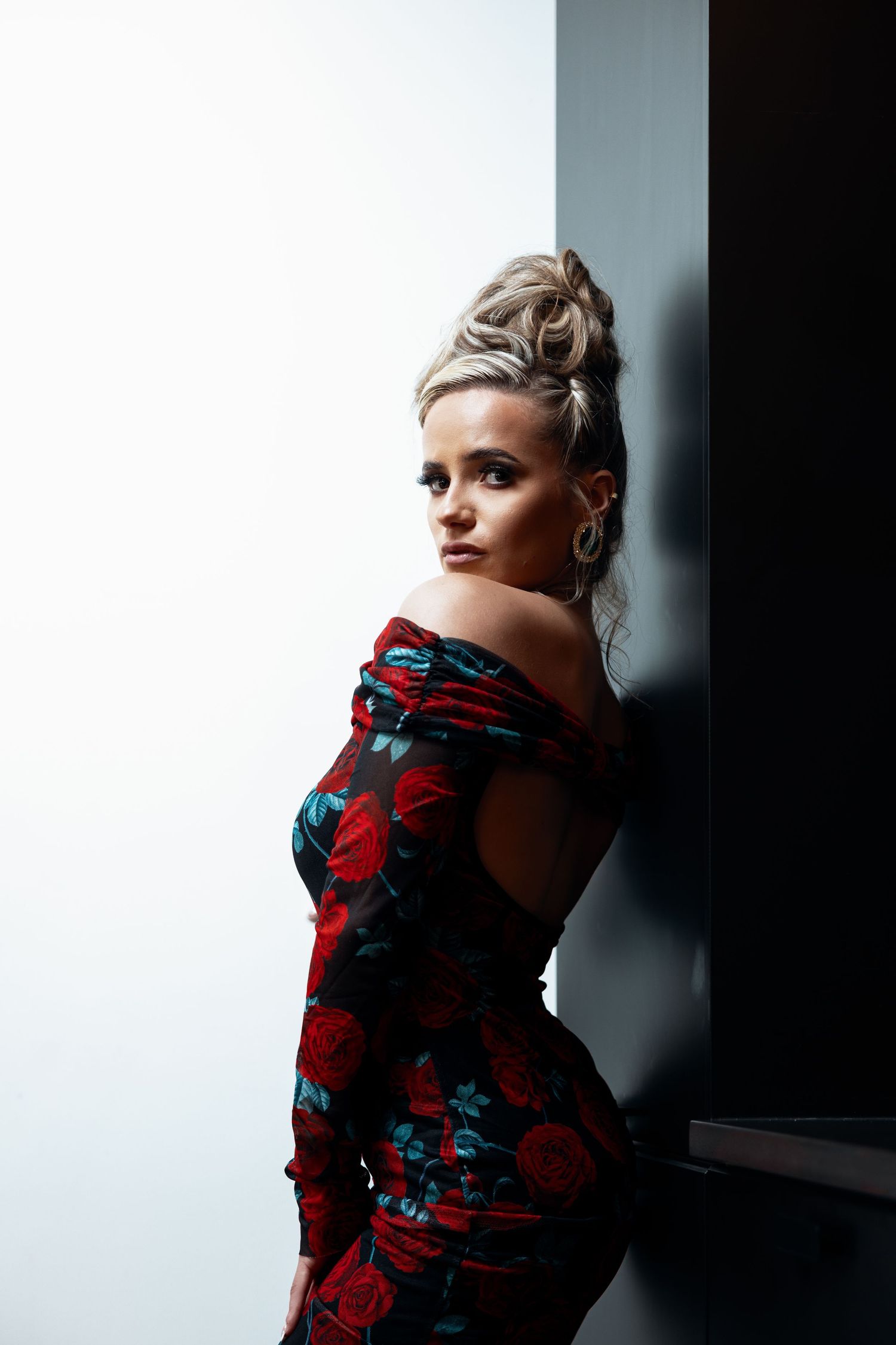 Woman in off-shoulder red floral dress with updo hairstyle posing against dark wall.