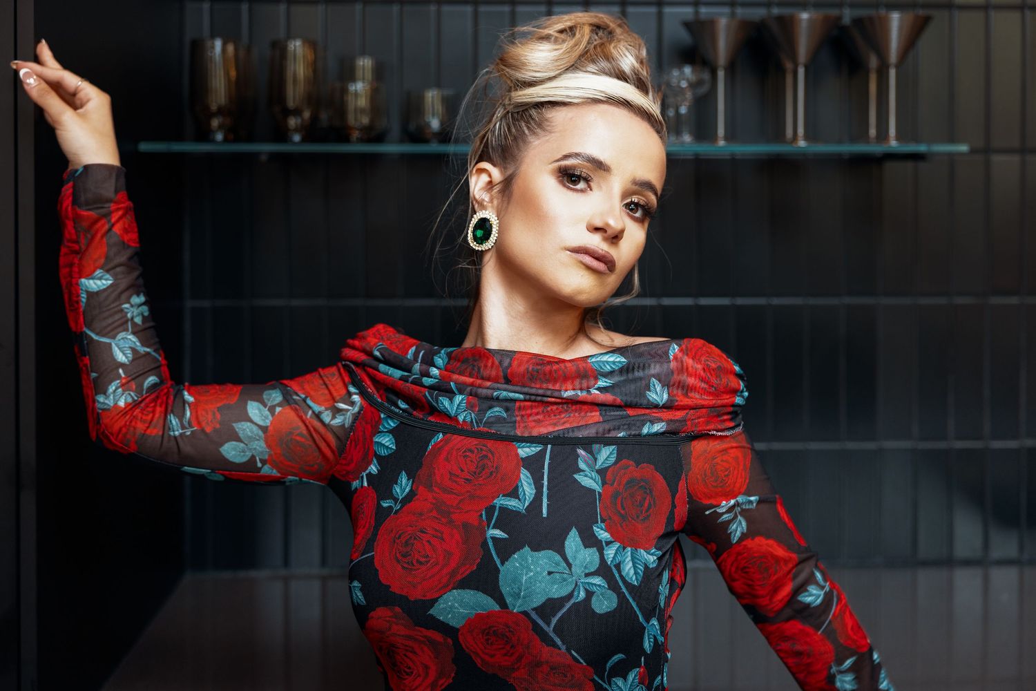 Young woman in red floral rose print dress with updo hairstyle posing against dark shelving.