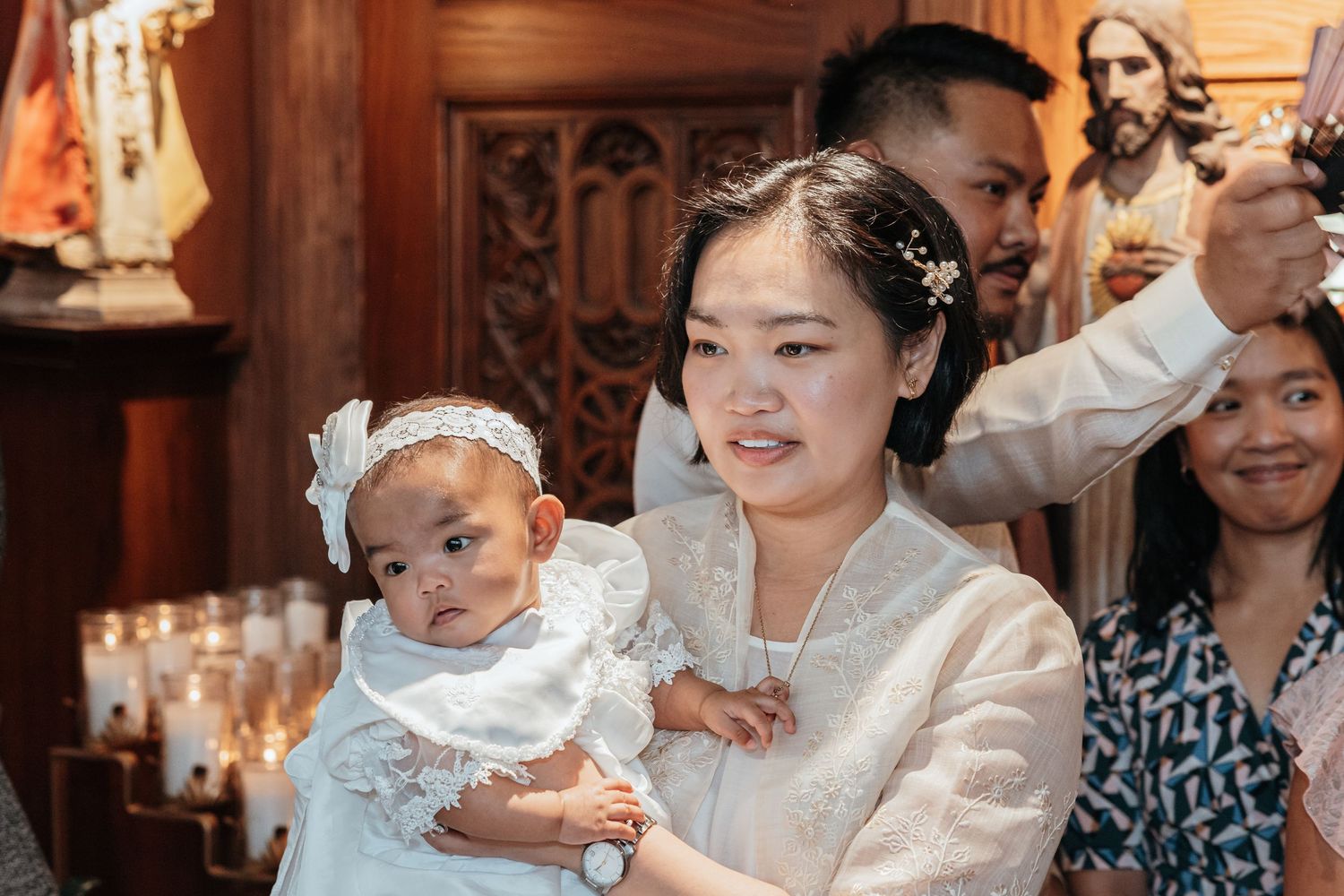 A smiling Filipino mother in white barong holds her baby in a christening gown near church candles.