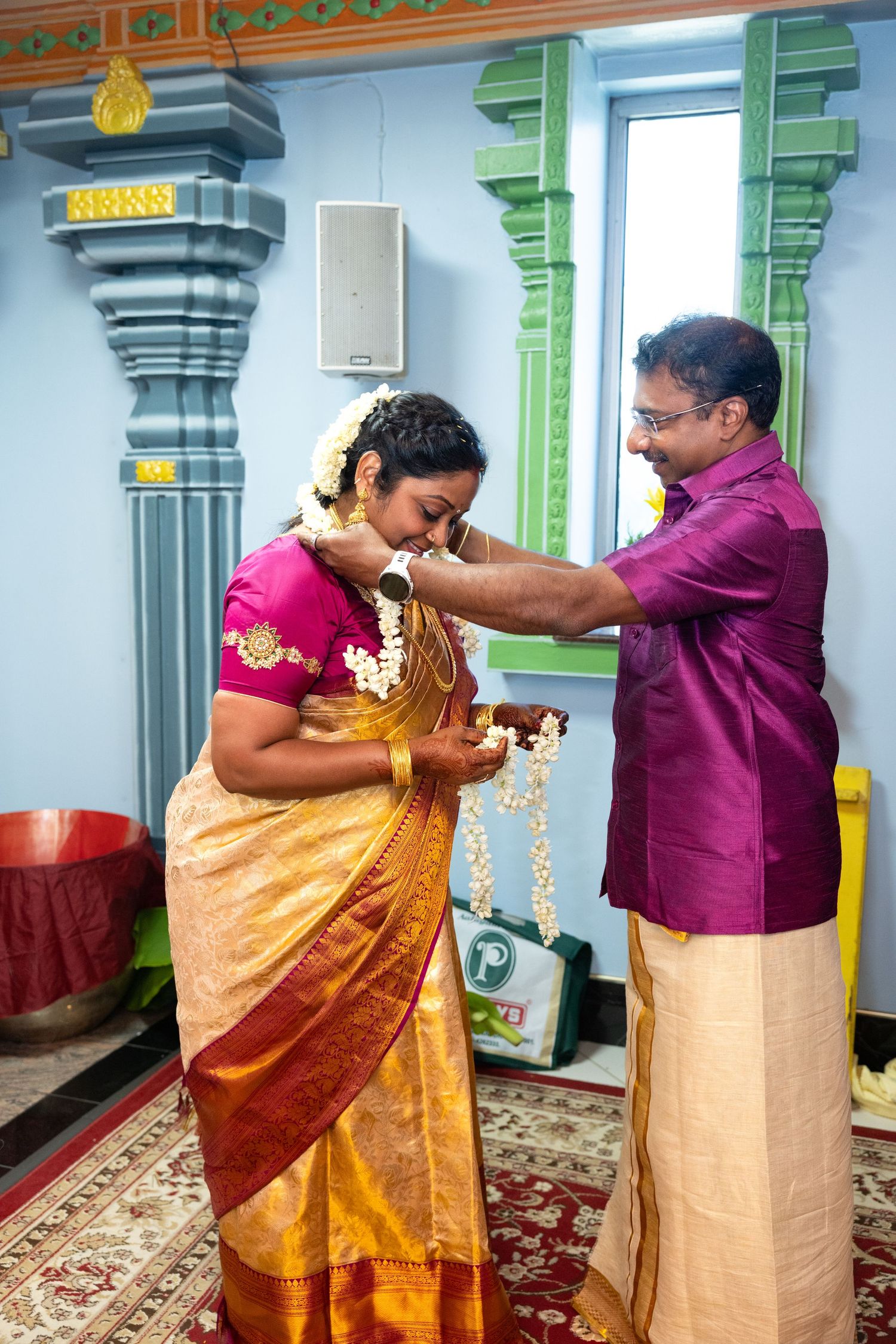 Man placing a flower garland on a woman in a golden silk saree during a Hindu ceremony.