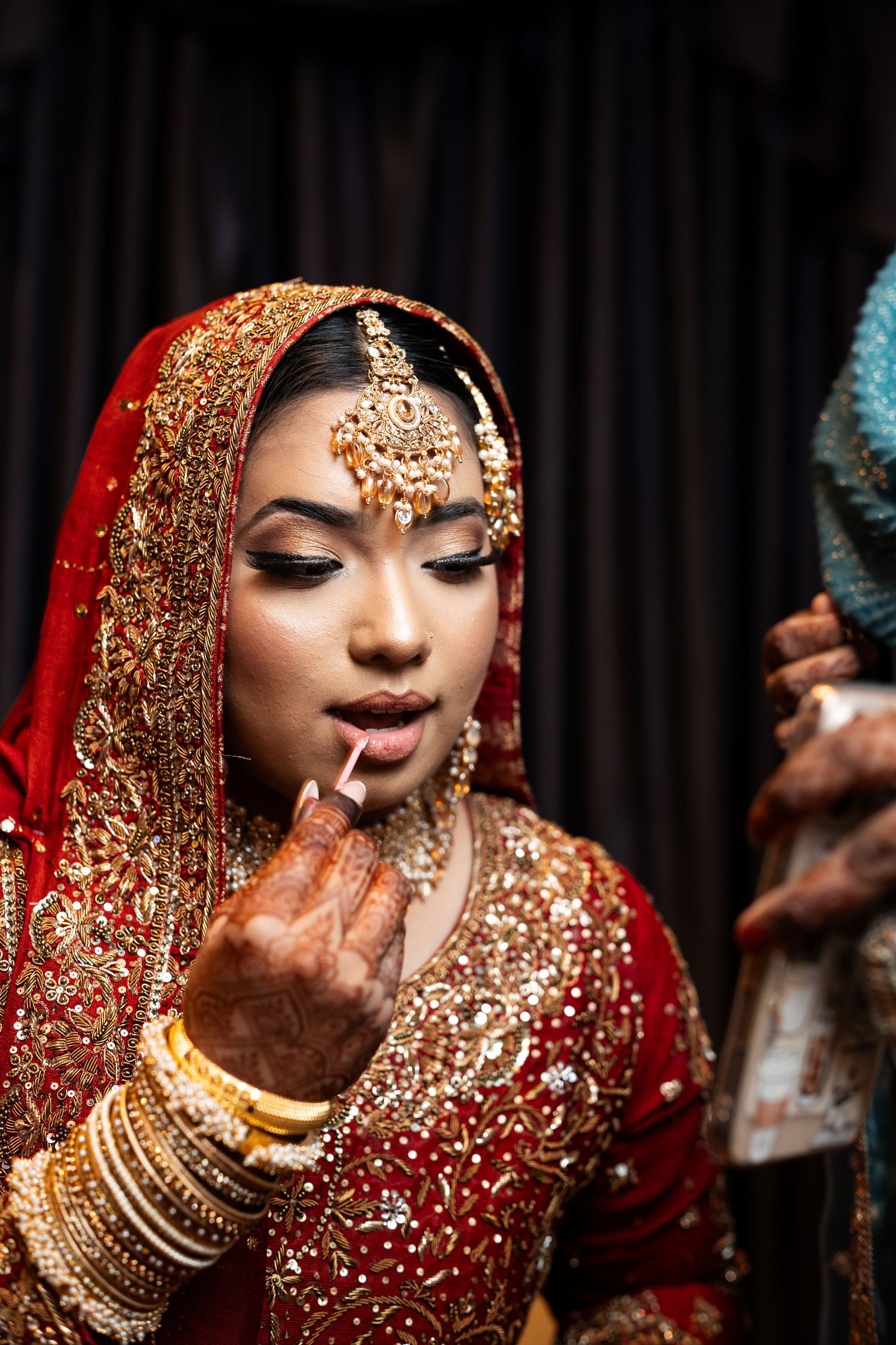 An Indian bride in ornate red and gold bridal wear applies lipstick while dressed in traditional wedding attire.