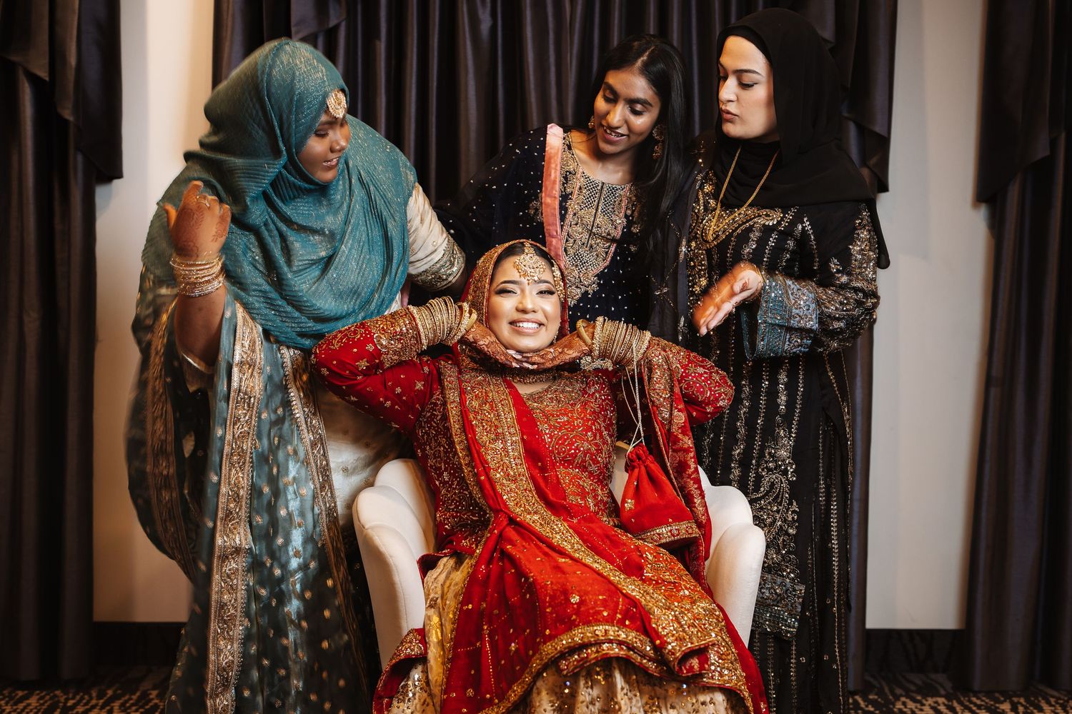 Bride in red bridal lehenga smiling broadly while three female attendants assist her preparation.